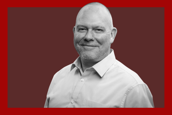 Scott Pawley: A middle-aged man with a bald head and beard, smiling, wearing a light-colored button-up shirt, against a dark red background with a red border.
