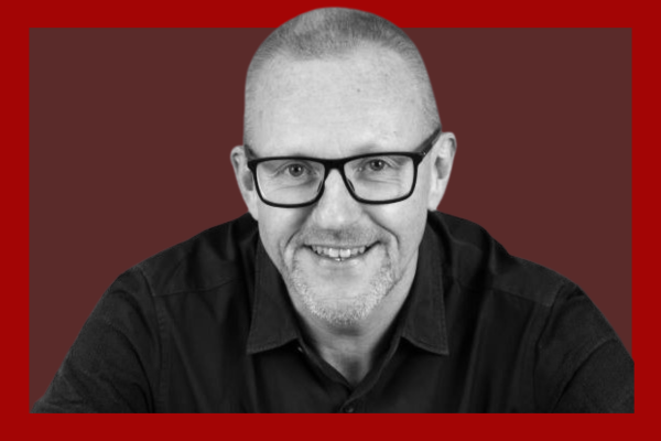 Clive Wratten: Smiling man with glasses, short hair, and a goatee wearing a black shirt, posed against a red background.