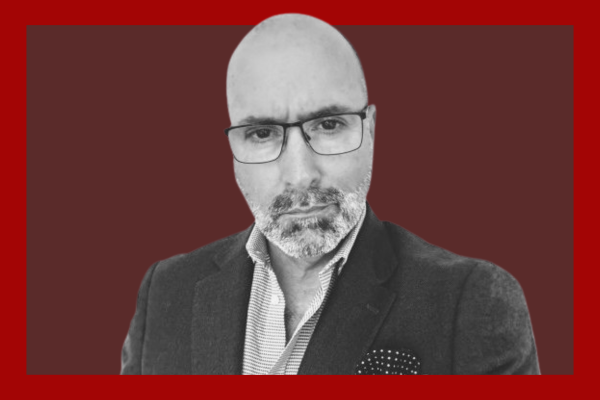 Paul Spencer: A man with glasses, a bald head, and a goatee, wearing a dark blazer and a collared shirt, taking a selfie against a dark red background.