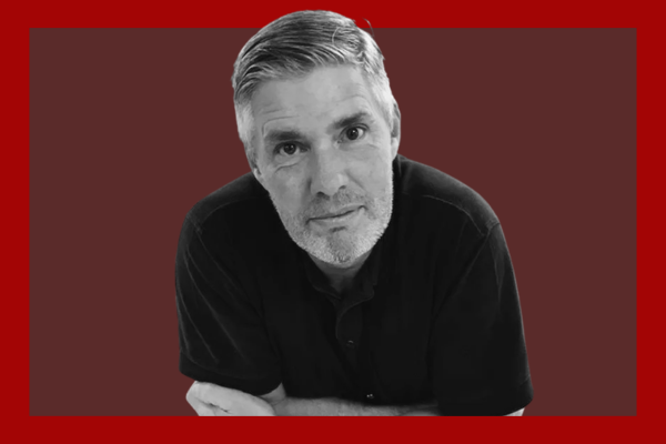 Neil Mason: Black and white portrait of a middle-aged man with gray hair and a beard, wearing a black polo shirt, against a red background.