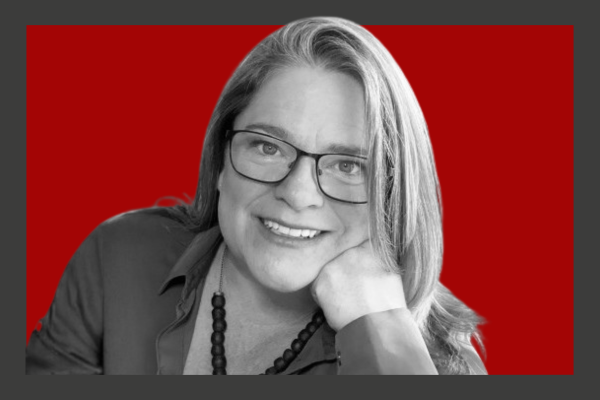 Joanna Nicholas: A woman with glasses smiling, resting her chin on her hand, against a red background.
