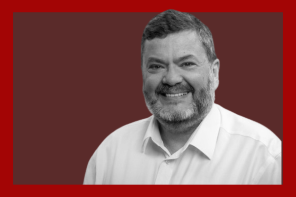 Mark Wilson: A smiling man with a beard and short hair wearing a white shirt, against a red and brown background.