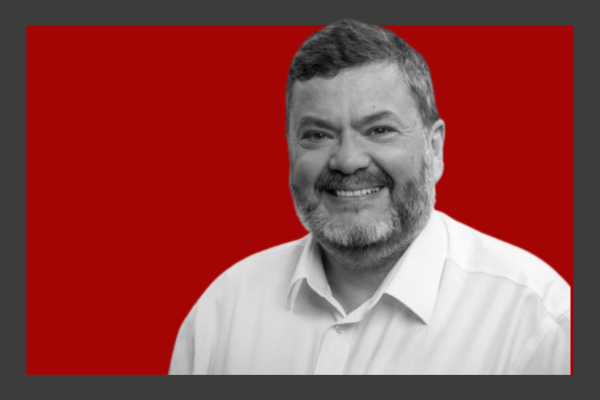 Mark Wilson: Black and white photo of a smiling man with a beard, wearing a white shirt, against a red background.