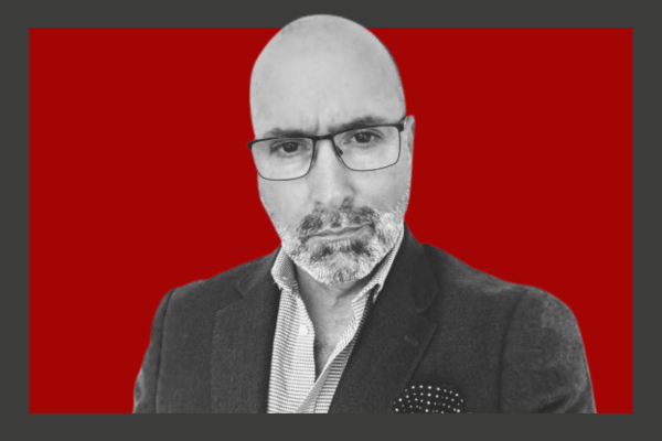 Paul Spencer: A monochrome photo of a bald man with glasses, wearing a blazer and collared shirt, against a red background.