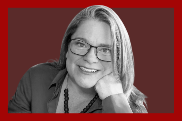 Joanna Nicholas: A woman with glasses, smiling, resting her chin on her hand, wearing a beaded necklace and a collared shirt, in a red and black background.
