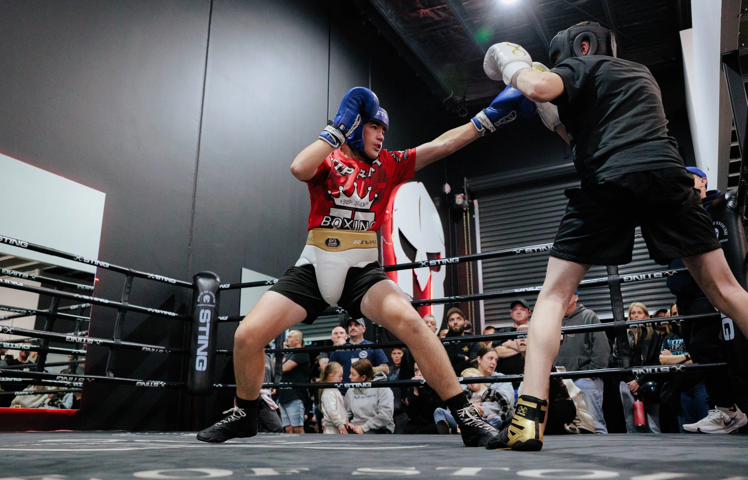 Two House of Stoush teen/adult members wearing boxing gloves, sparring helmets, and athletic clothing engaging in a sparring match inside a boxing ring surrounded by community spectators.