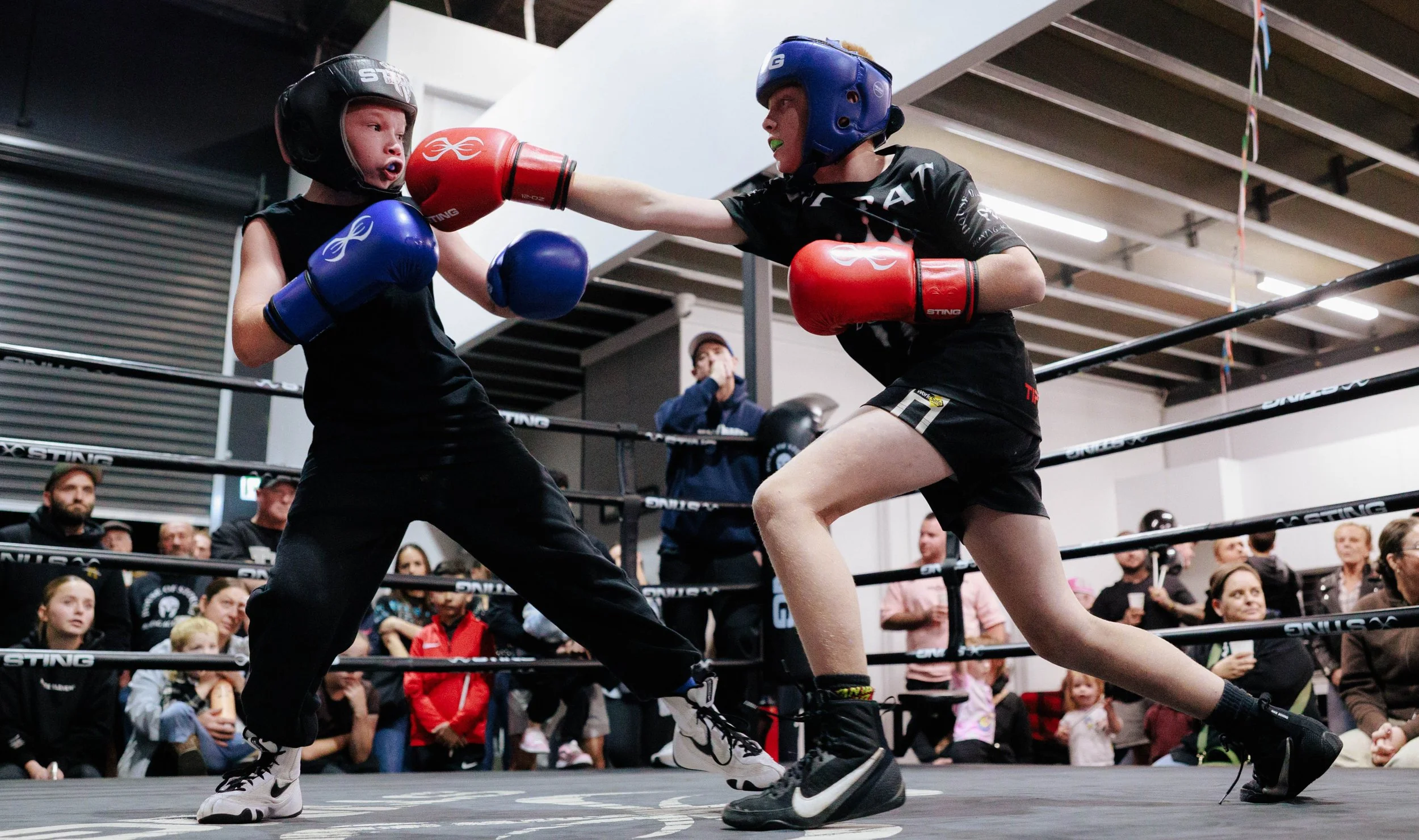 Two young House of Stoush kid members wearing sparring helmets, boxing gloves, and athletic shorts engaging in a sparring match inside a ring, surrounded by community spectators.