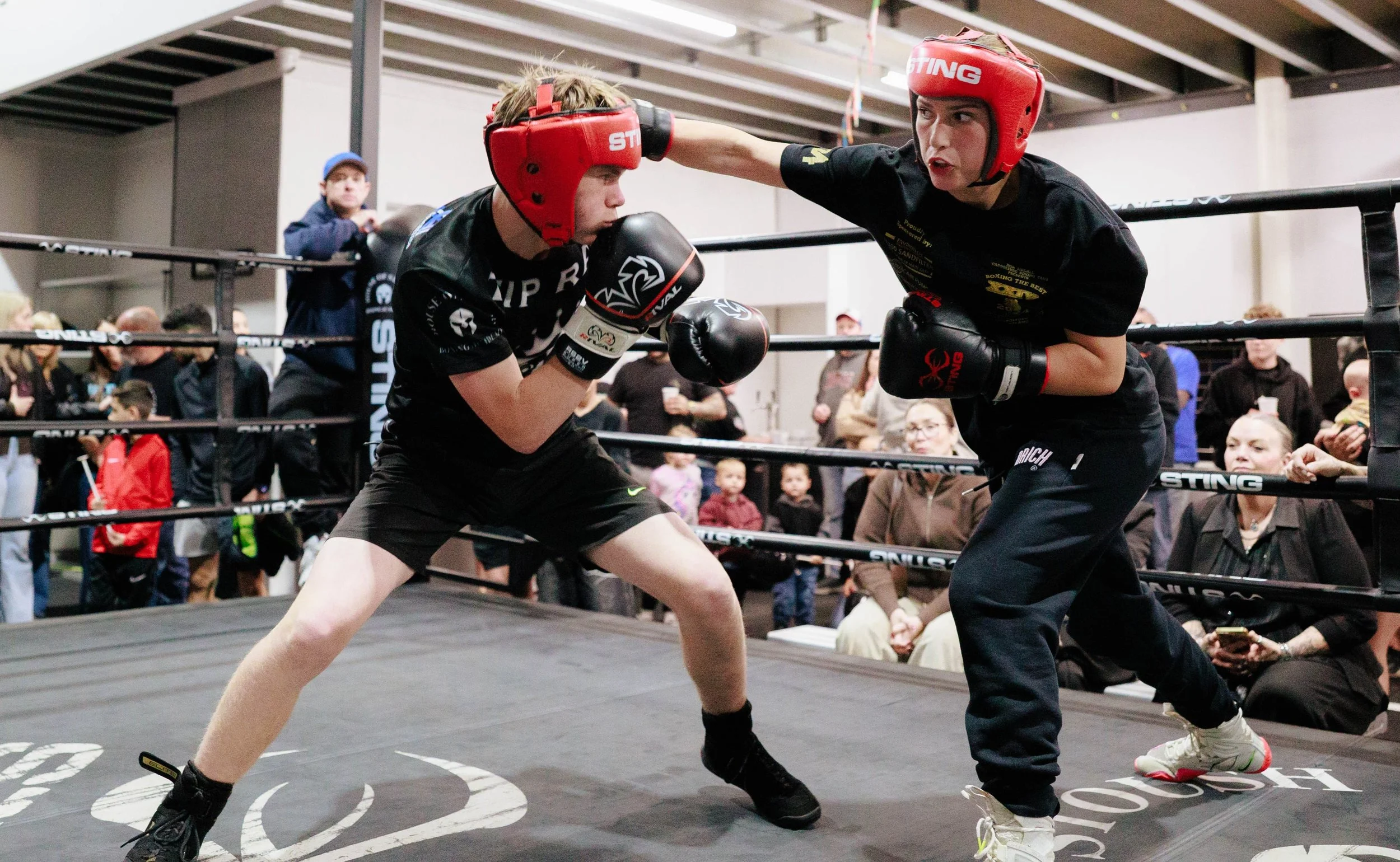 Two House of Stoush teen/adult members wearing sparring helmets, boxing gloves and House of Stoush merchandise engaging in a sparring match inside a boxing ring, surrounded by community spectators.