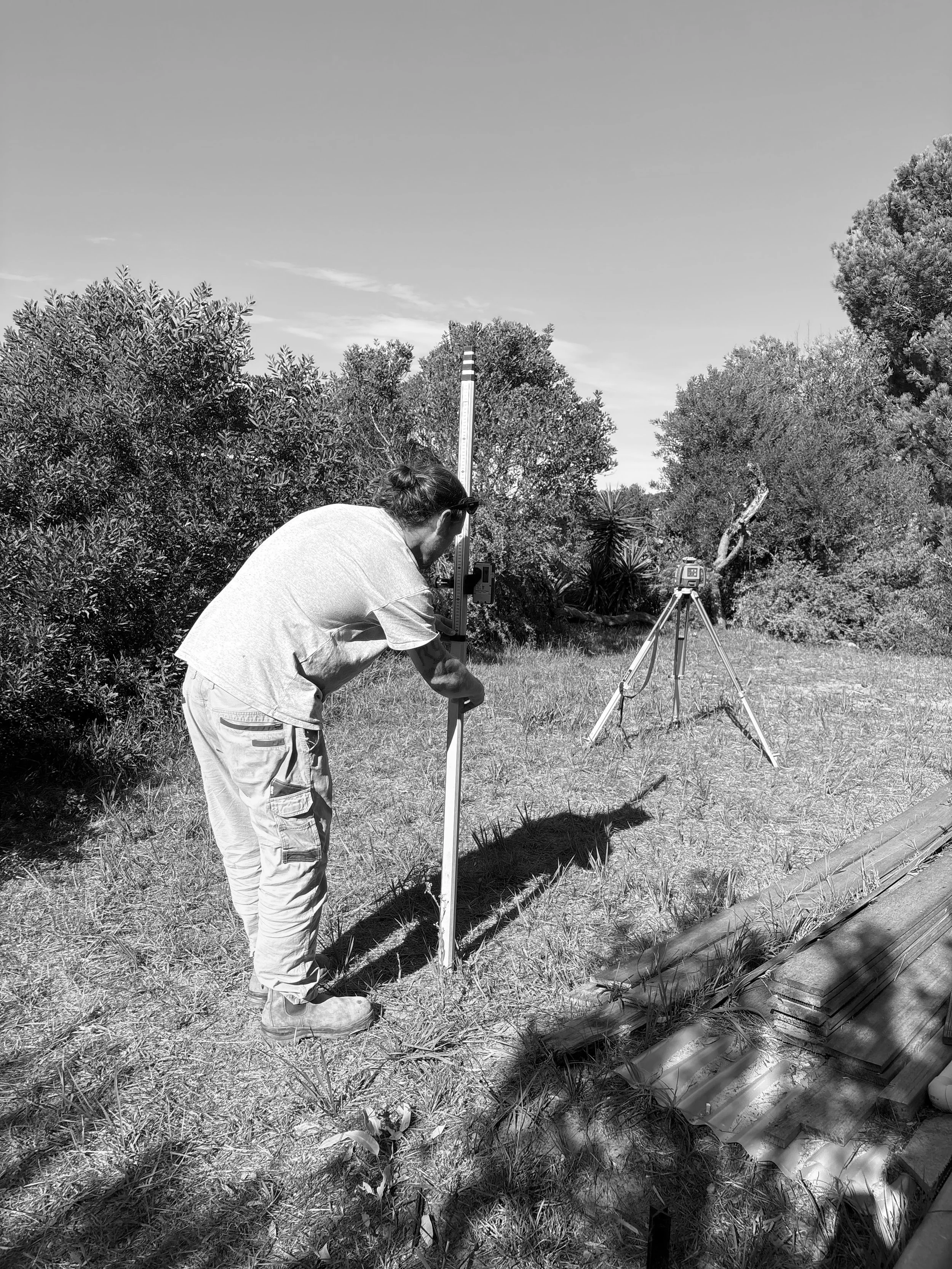 A person in a white shirt and light-colored pants is operating surveying equipment outdoors, surrounded by trees and grass, with a clear sky overhead.