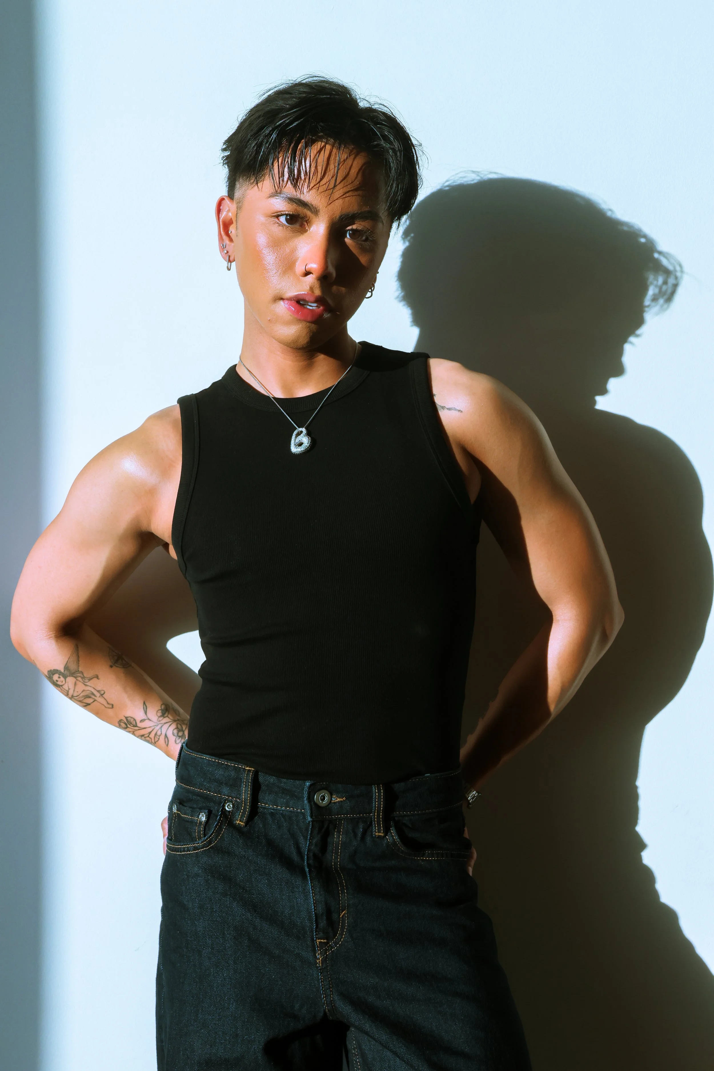 A woman with short dark hair and multiple tattoos wears a black tank top and dark jeans, standing against a white wall with a shadow cast behind her.