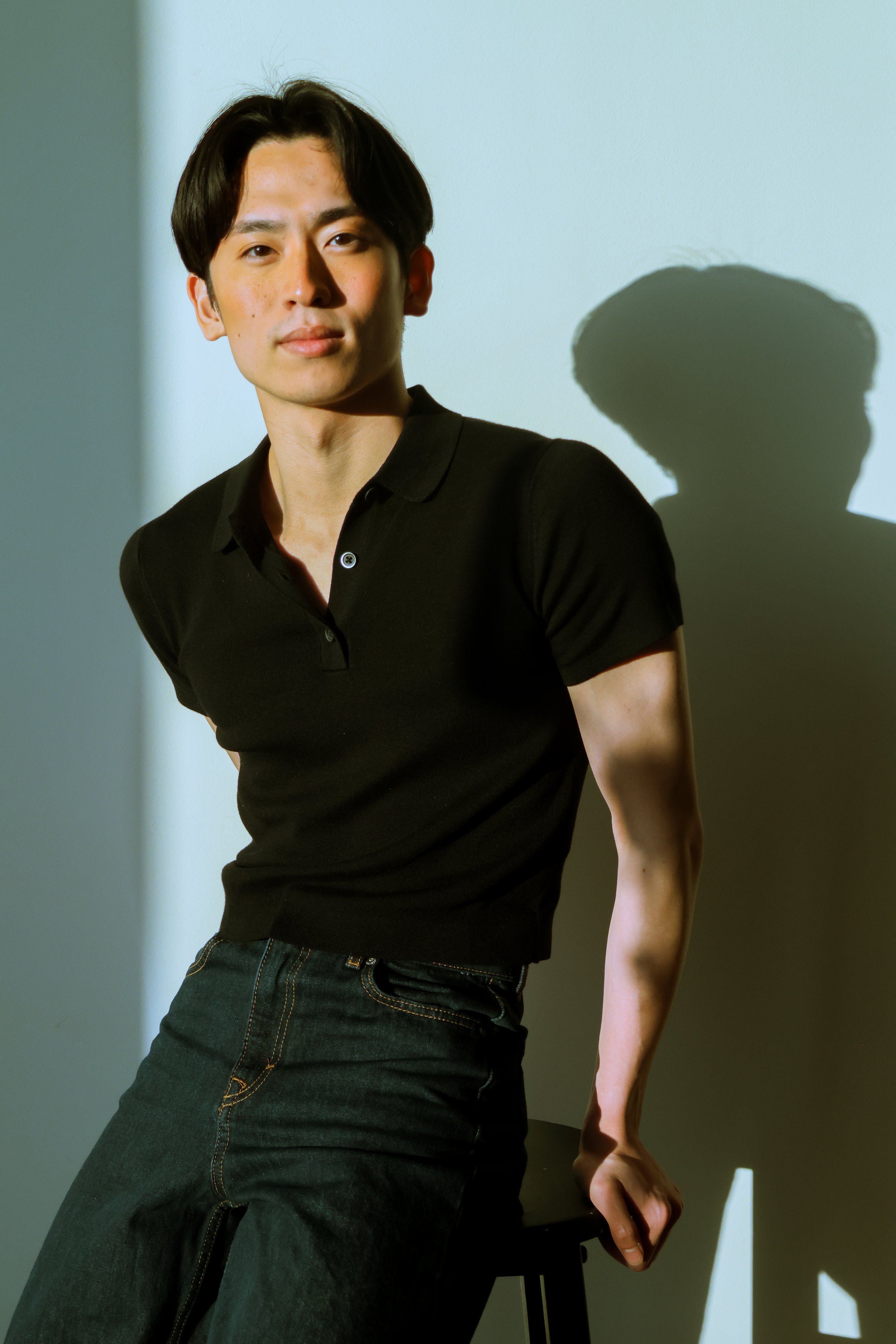 A young man with dark hair, wearing a black polo shirt and dark jeans, sits on a stool in front of a light-colored wall. His shadow is cast onto the wall to his right.