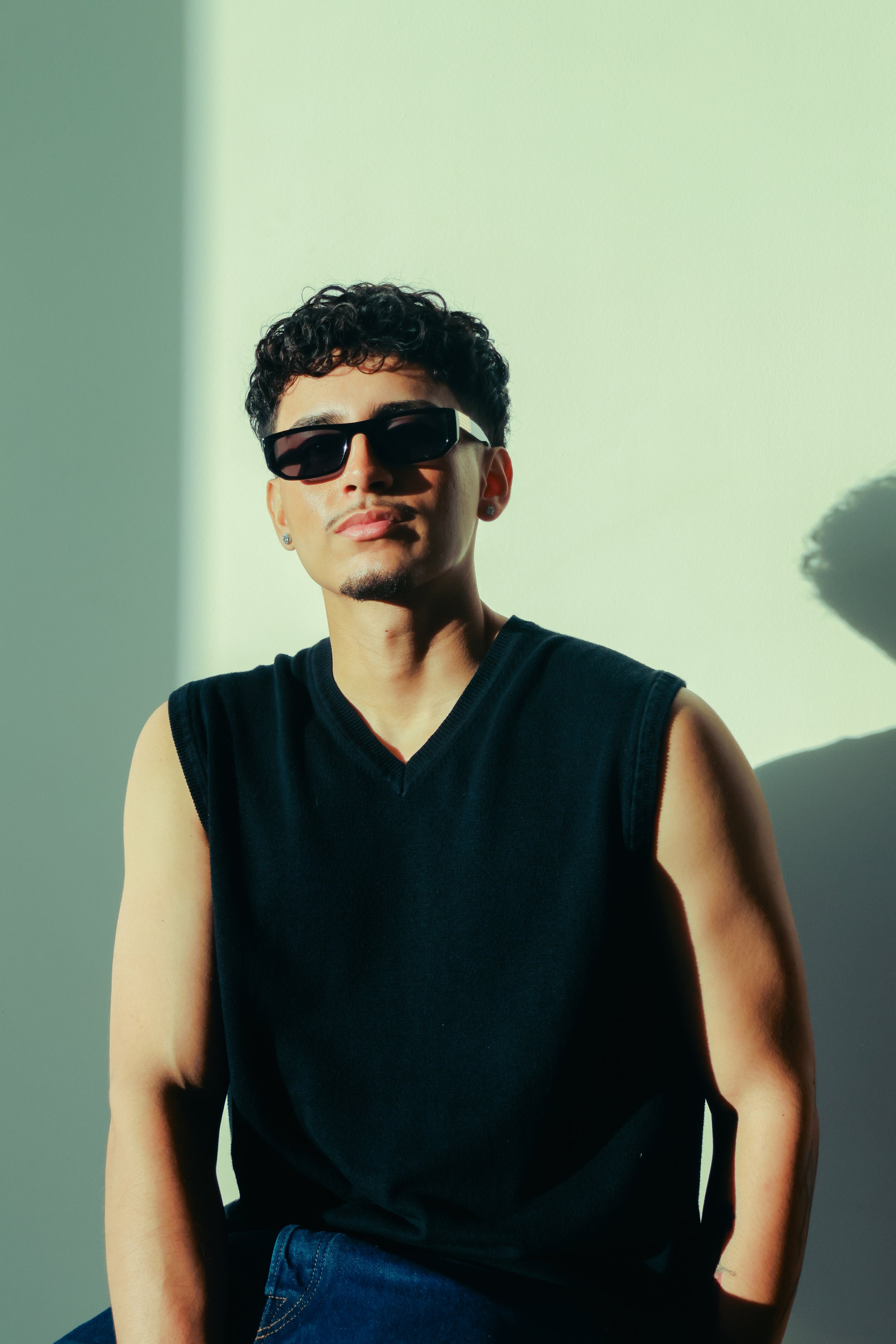 A young man with curly dark hair wearing black sunglasses, a sleeveless black shirt, and earrings, sitting in front of a plain wall with sunlight casting a shadow.