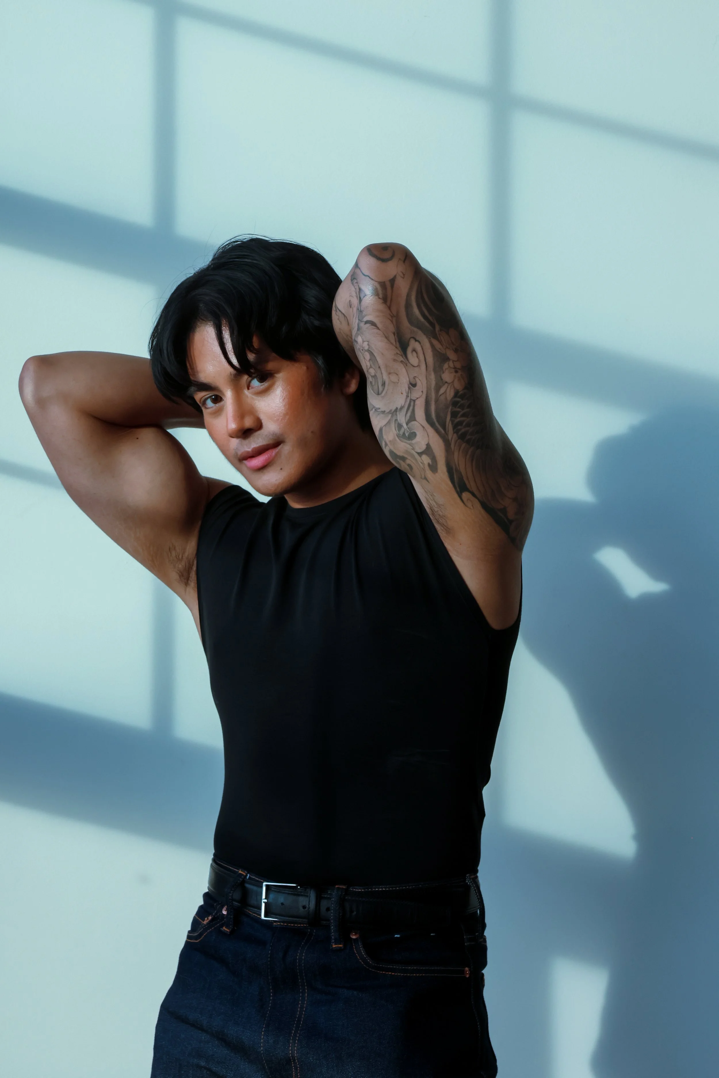 A young man with black hair and tattoos on his arm, wearing a sleeveless black shirt and dark jeans, standing against a light-colored wall with shadow patterns.