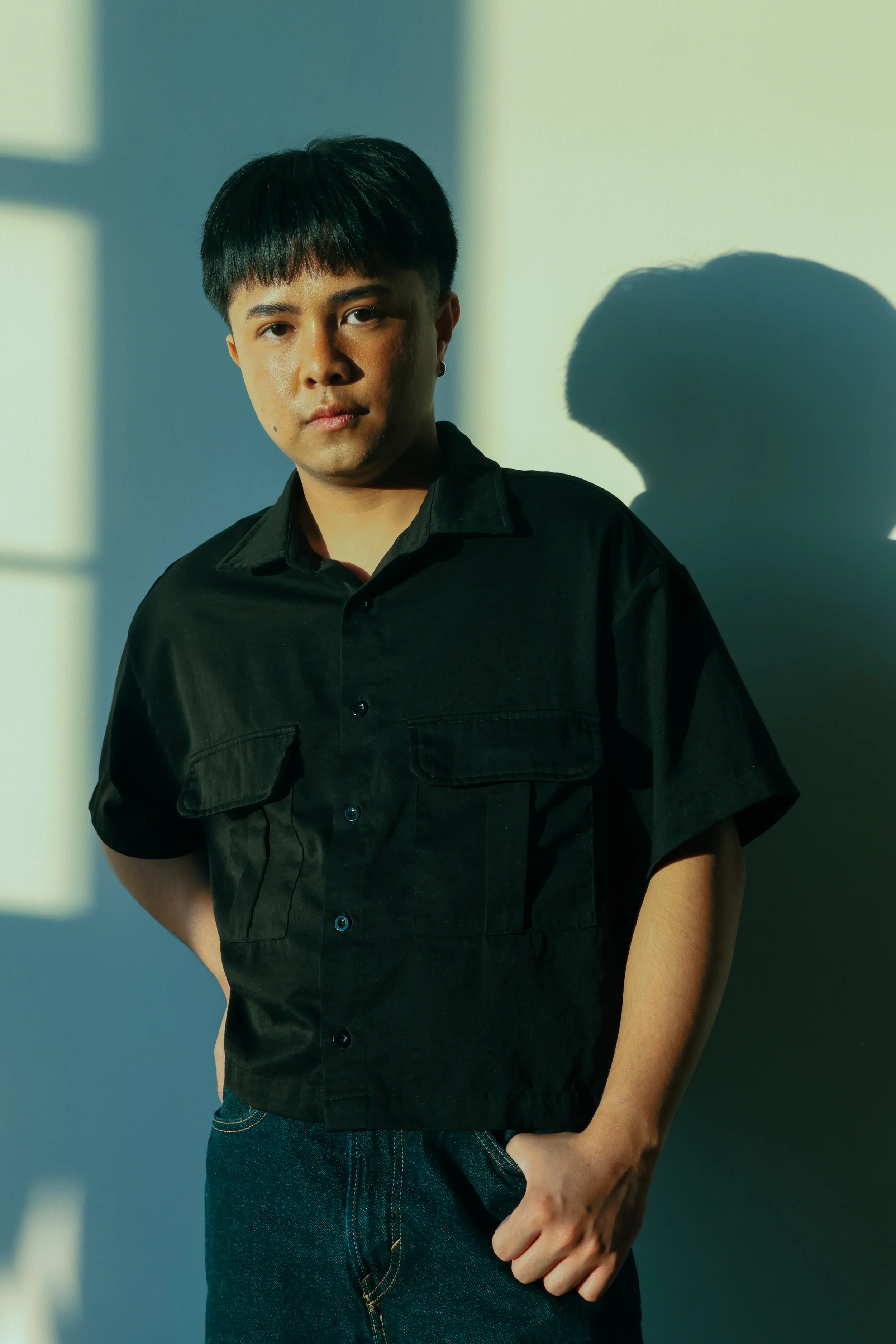 A young person with short dark hair and earrings, wearing a black button-up shirt and dark jeans, standing indoors with sunlight casting shadows on the wall behind them.