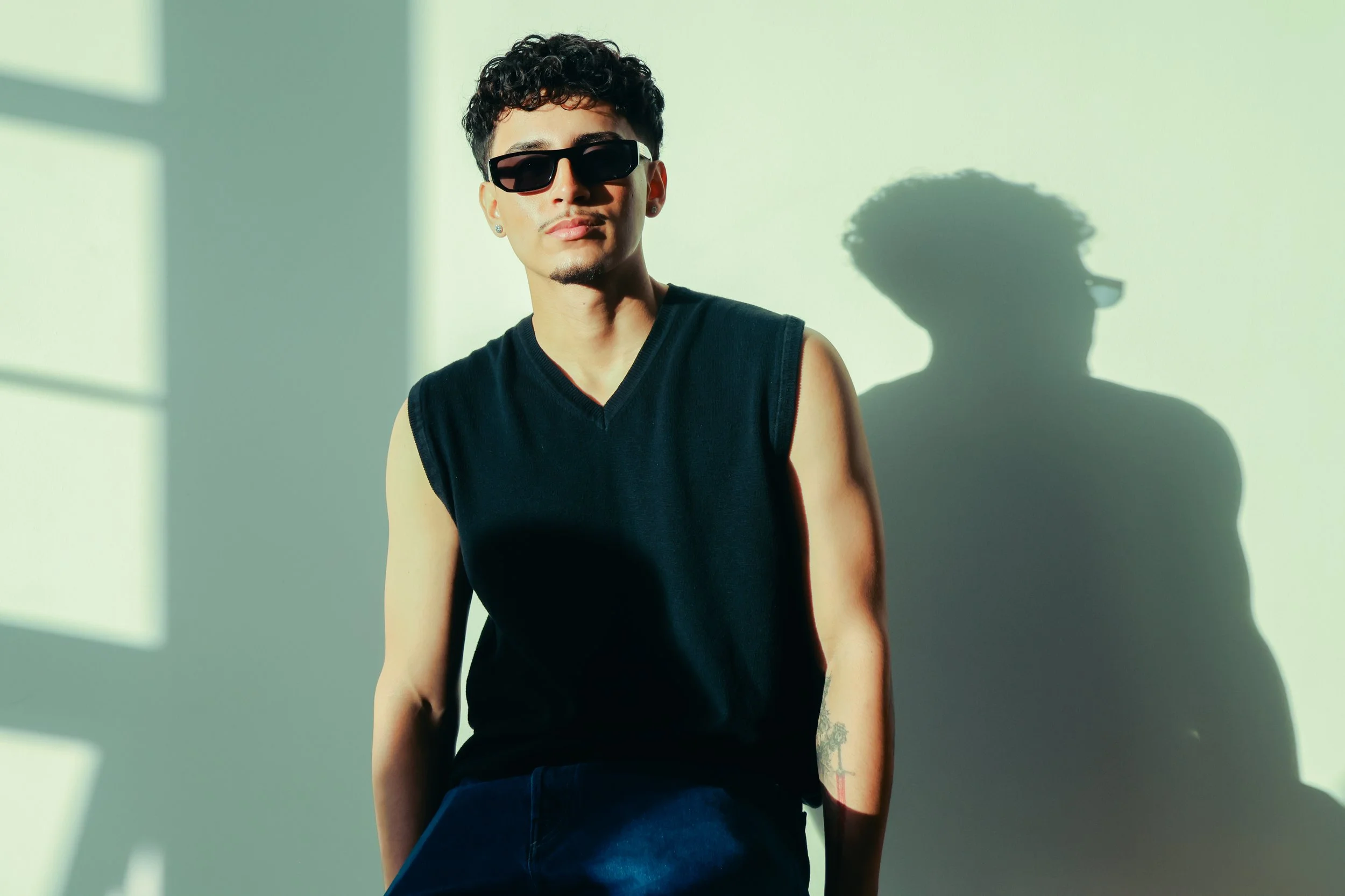 A young man with curly hair, wearing sunglasses and a sleeveless black shirt, standing in front of a light-colored wall with sunlight creating a shadow of him on the wall.
