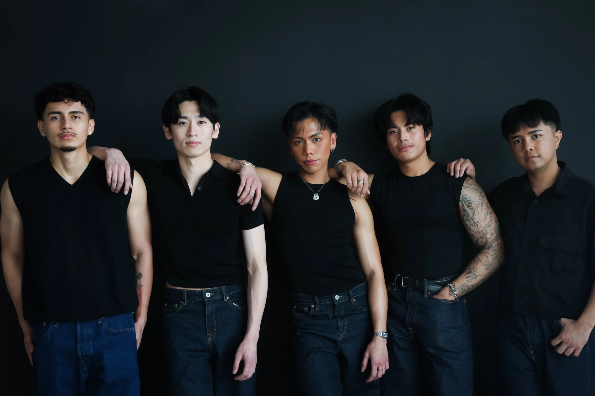 Five young adults standing side by side against a dark background, wearing black clothing, with arms around each other's shoulders, looking at the camera.