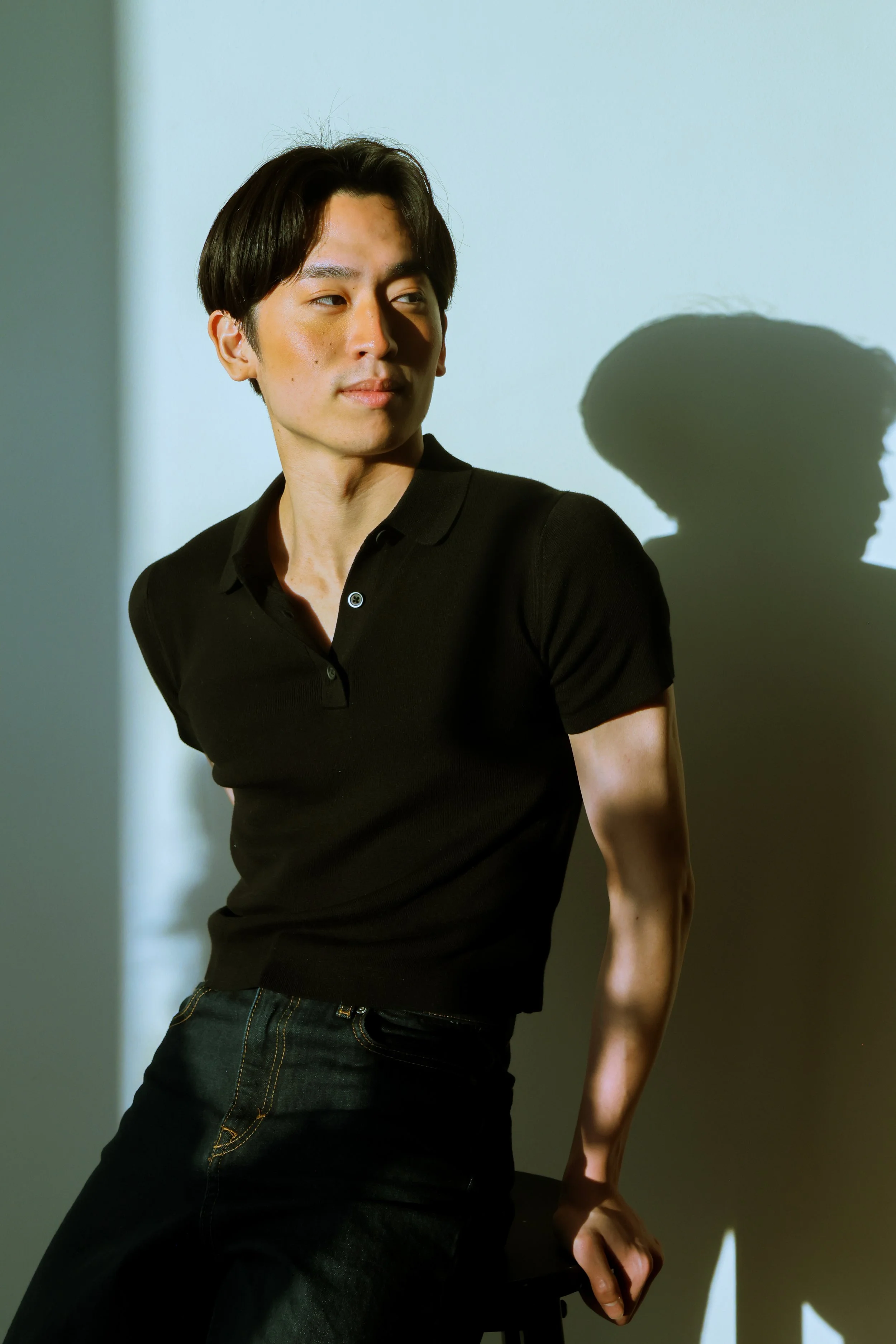 A young man with dark hair wearing a black polo shirt and dark jeans, sitting on a stool against a light-colored wall, with a shadow cast on the wall behind him.