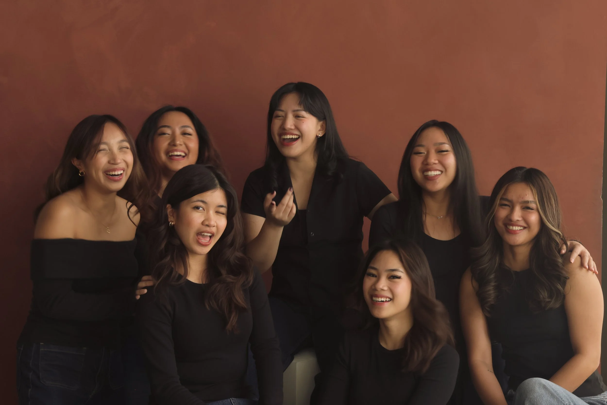 A group of nine young women, all wearing black tops, laughing and smiling together against a warm brown background.