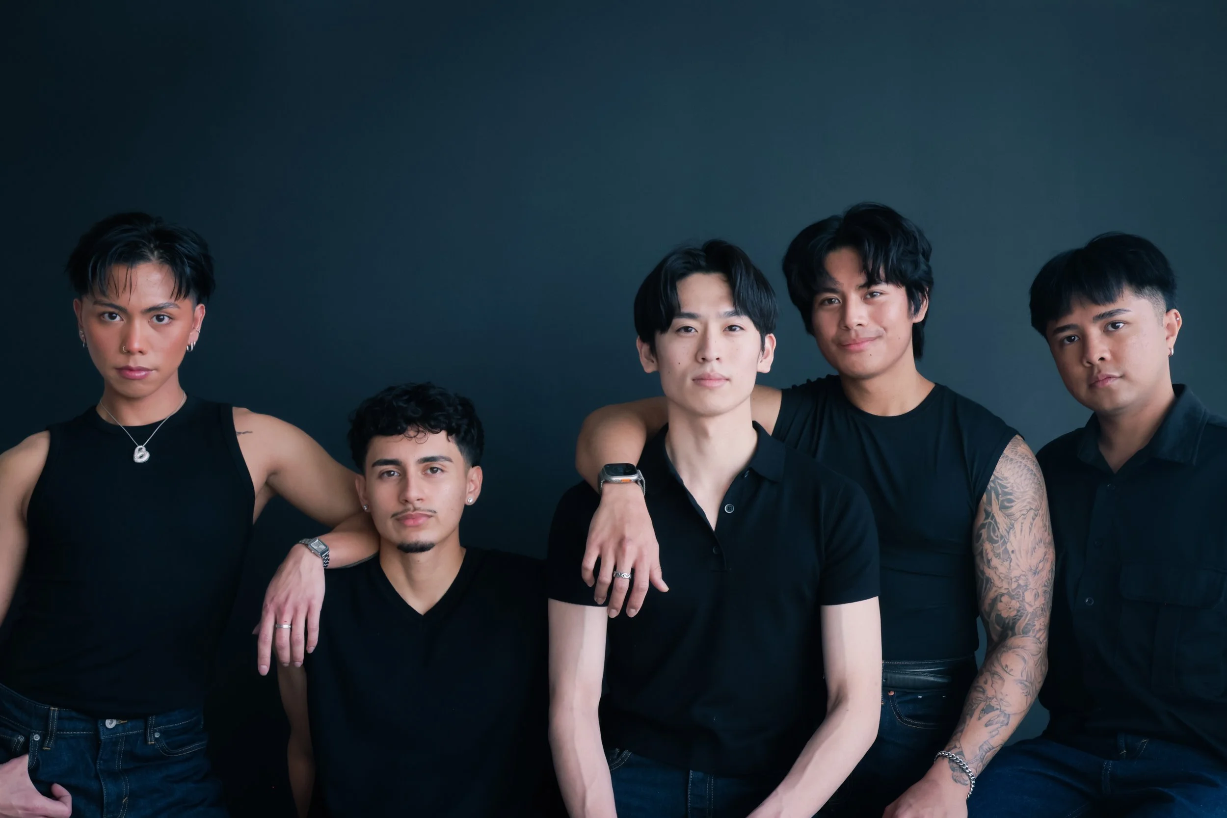 Group of five young men standing against a dark background, wearing black clothing and looking at the camera.