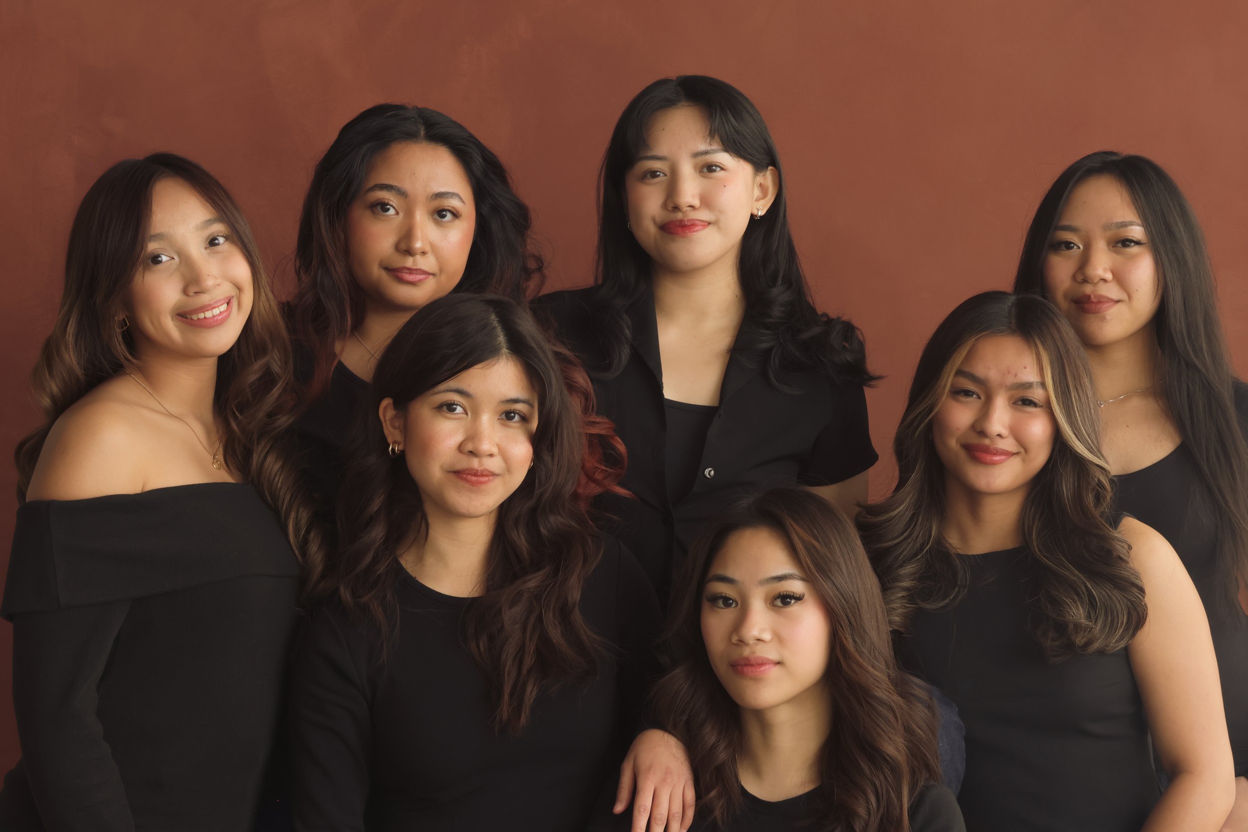 Group of eight young women standing together against a brown background, dressed in black tops, smiling and looking at the camera.