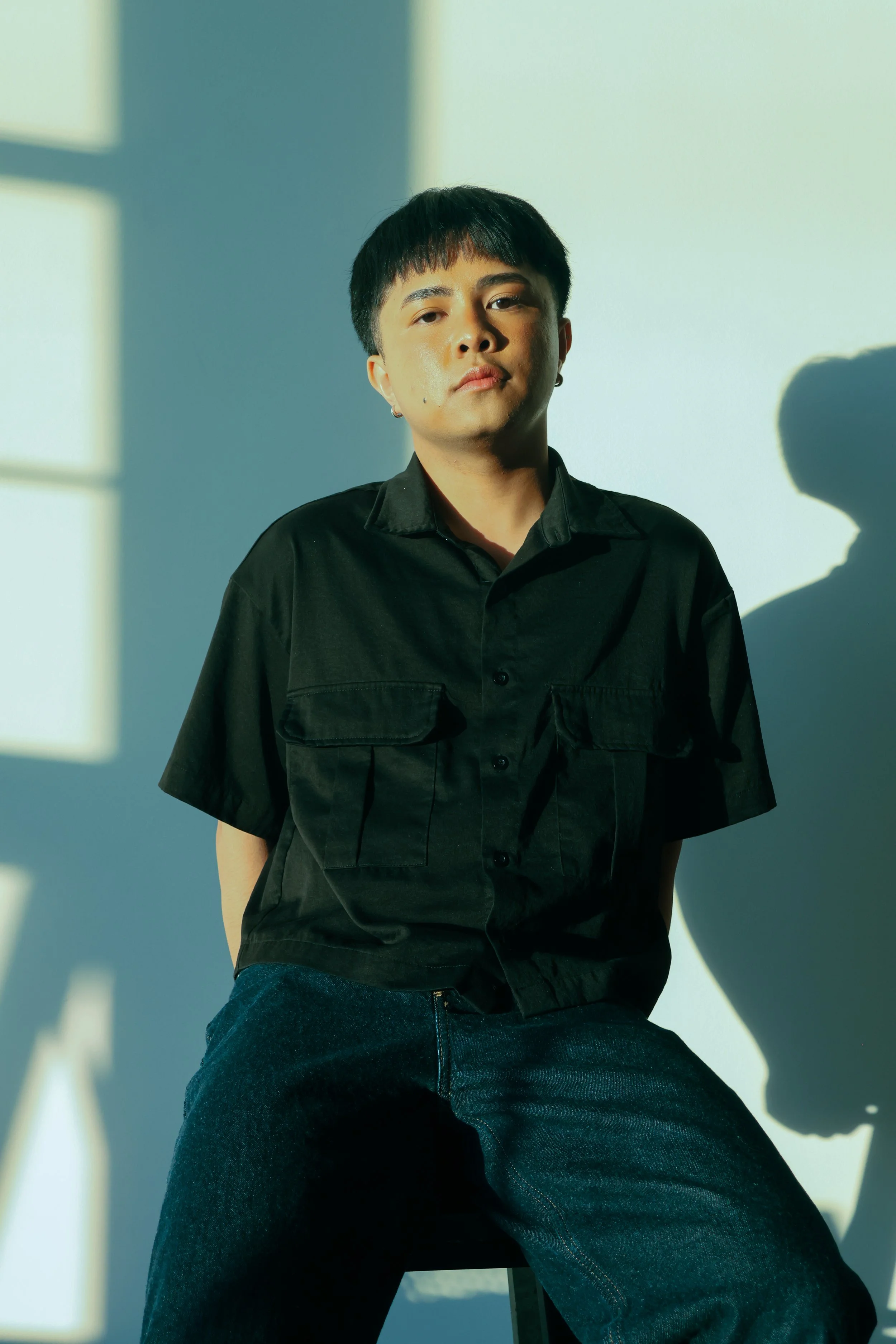 Person with short dark hair wearing a black shirt sitting in front of a wall with sunlight creating shadows.