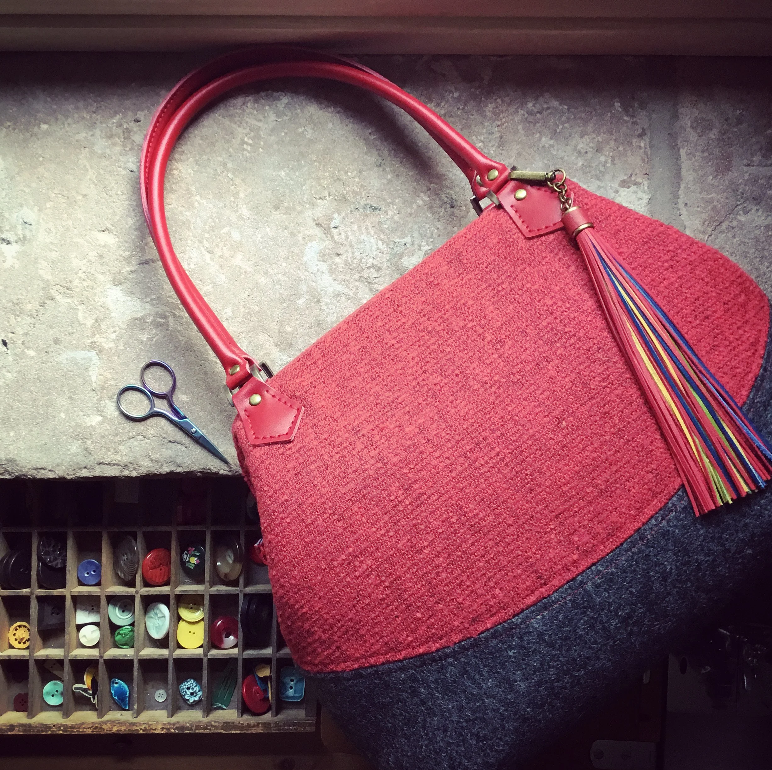 A red and gray handbag with a colorful tassel, placed on a work surface next to a pair of scissors and a button organizer.