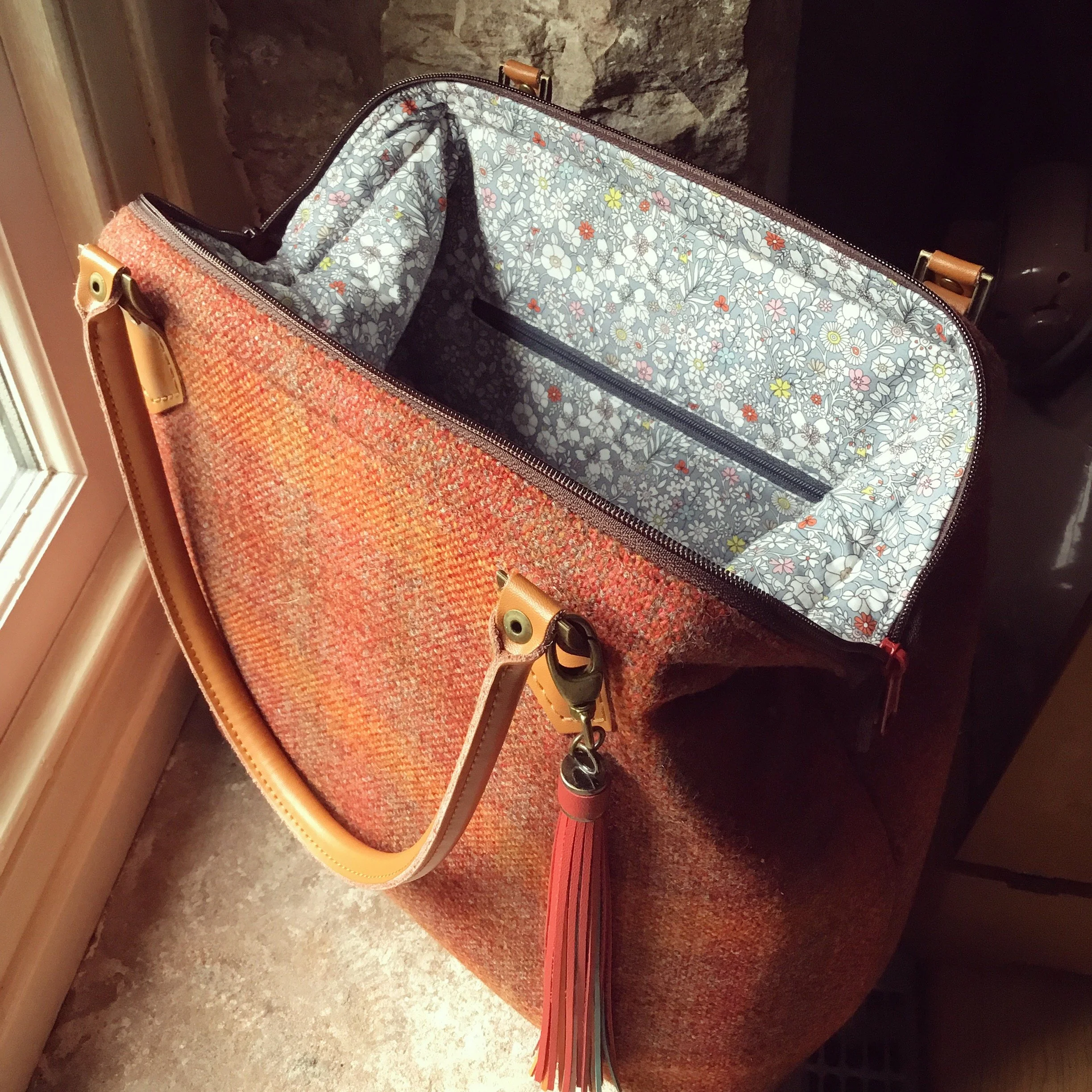 Open tote bag with floral patterned interior lining and leather handles resting on a textured surface near a window.