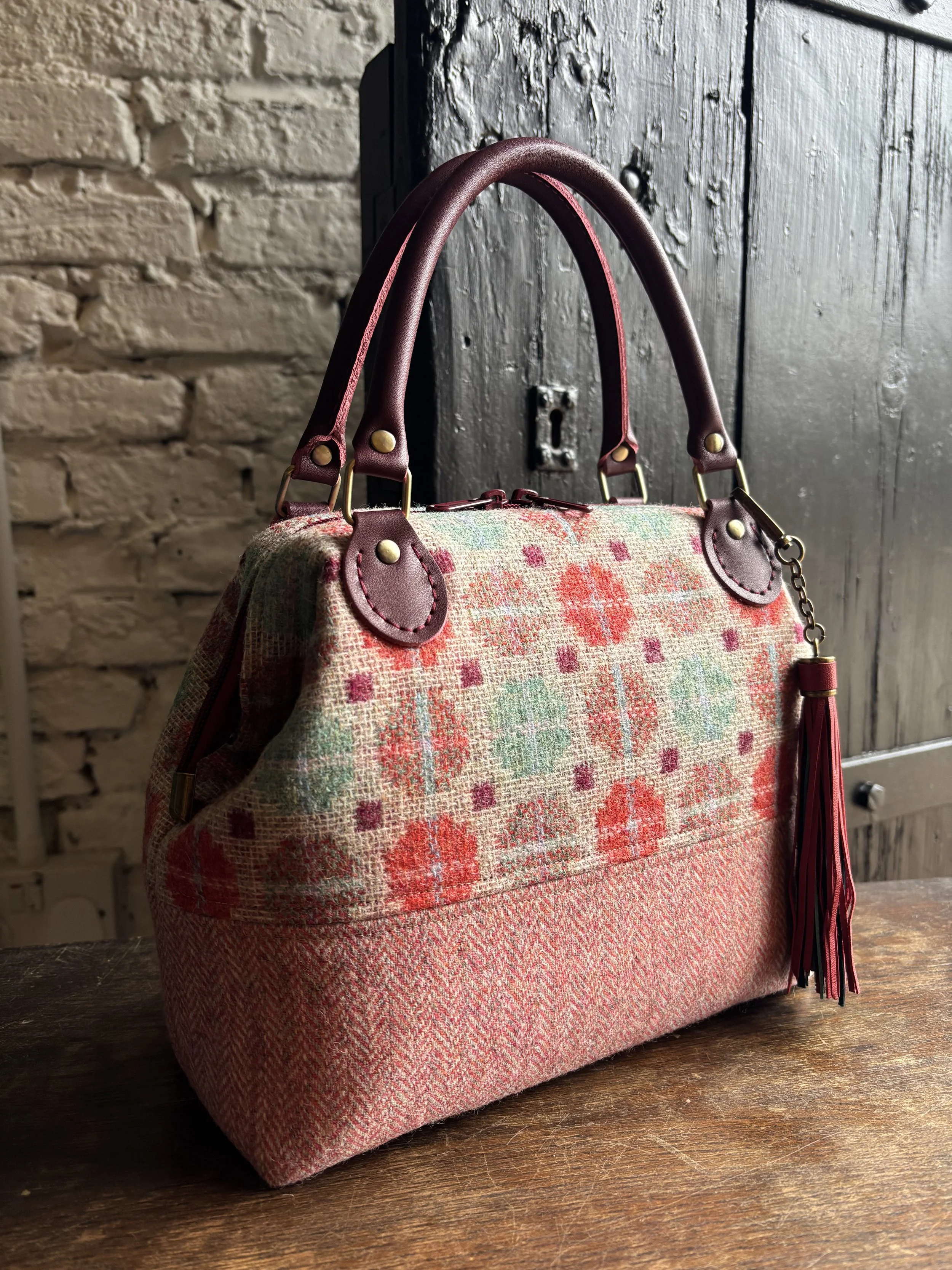 A stylish handbag with pink, red, and green floral and geometric patterns, featuring leather handles, a leather tassel charm, and brass hardware, resting on a wooden surface with a rustic brick and wood-paneled background.