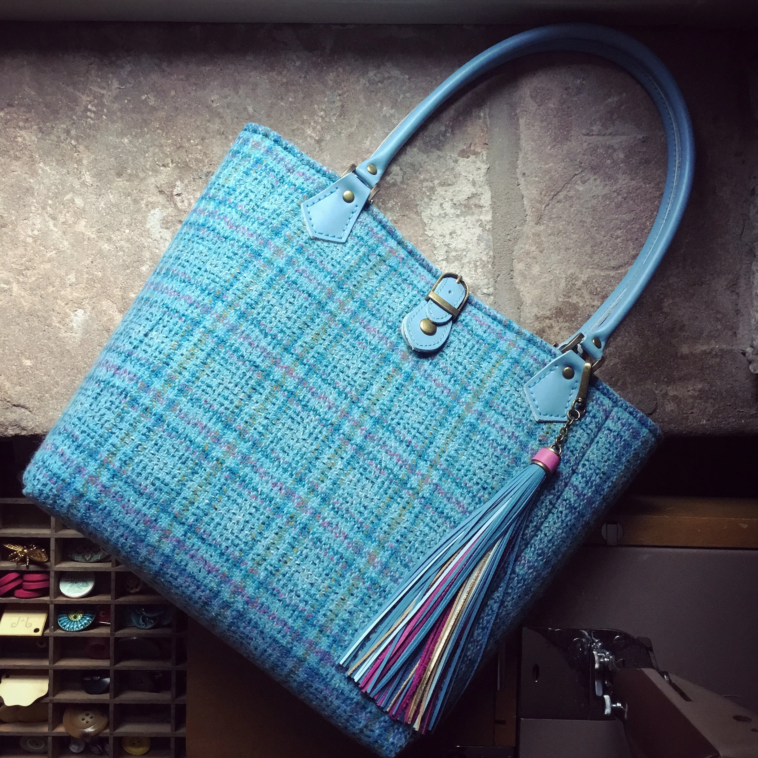 Blue plaid tote bag with light blue handles and multicolored fringe tassel hanging from a chain on one handle, resting on a stone surface.