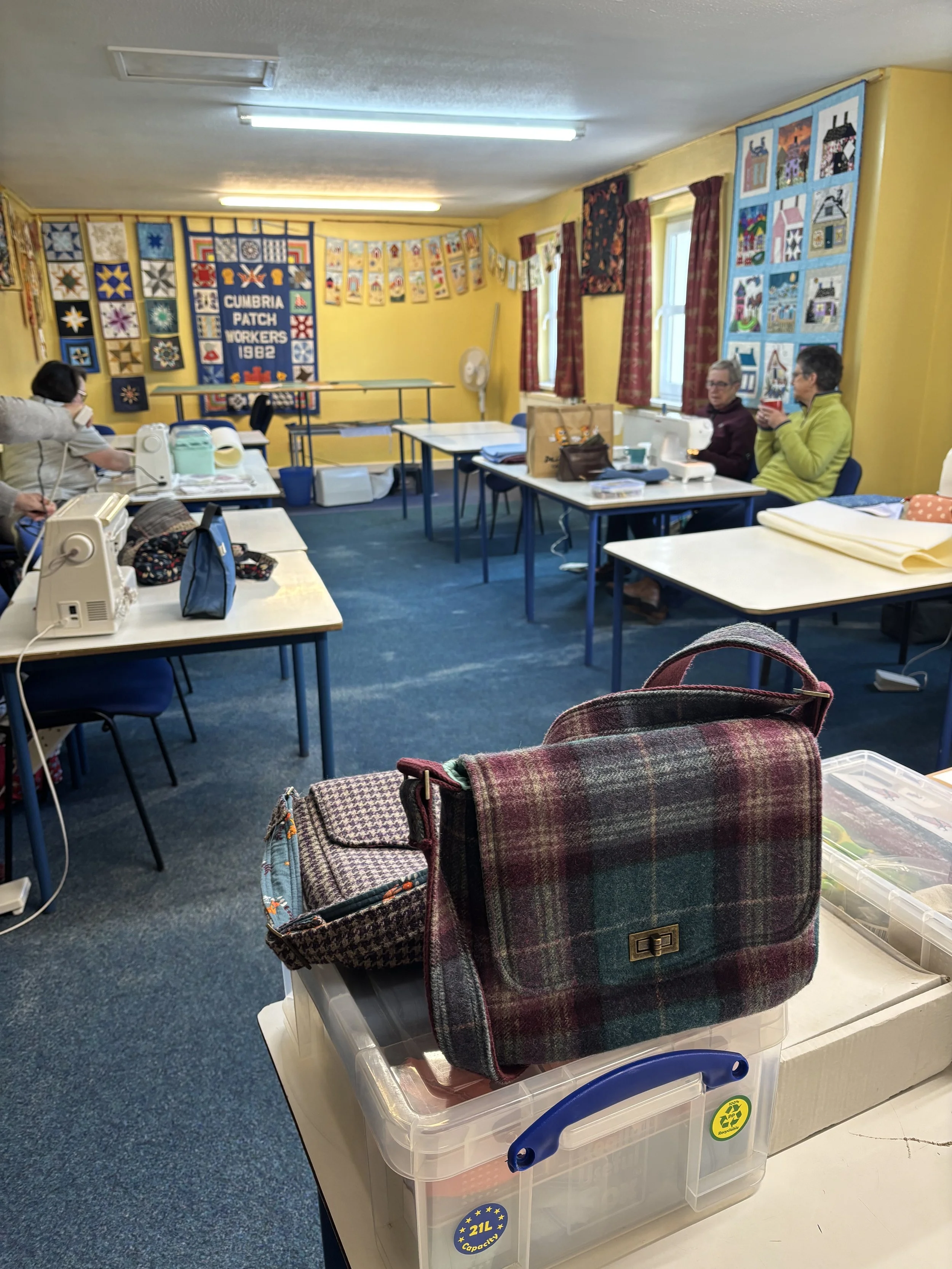 A quilting or craft workshop room with yellow walls, decorated with quilts and bunting. Several tables with sewing and craft supplies are arranged, with a few women working or conversing. Quilts are displayed on the walls and windows, and a banner reads 'CUMBRIA PATCH WORKERS 1982'.