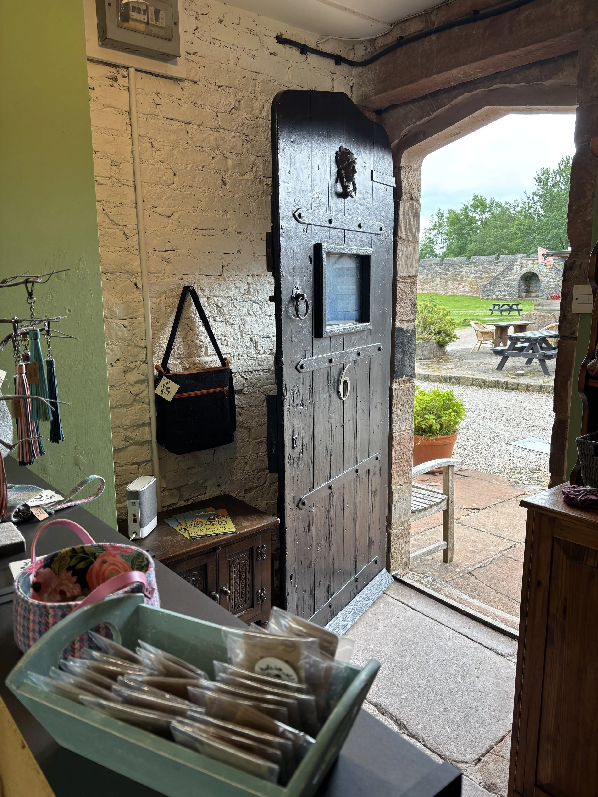 Open black wooden door with small window and metal hardware at the entrance of a rustic building, overlooking a courtyard with outdoor seating and a stone wall in the background.