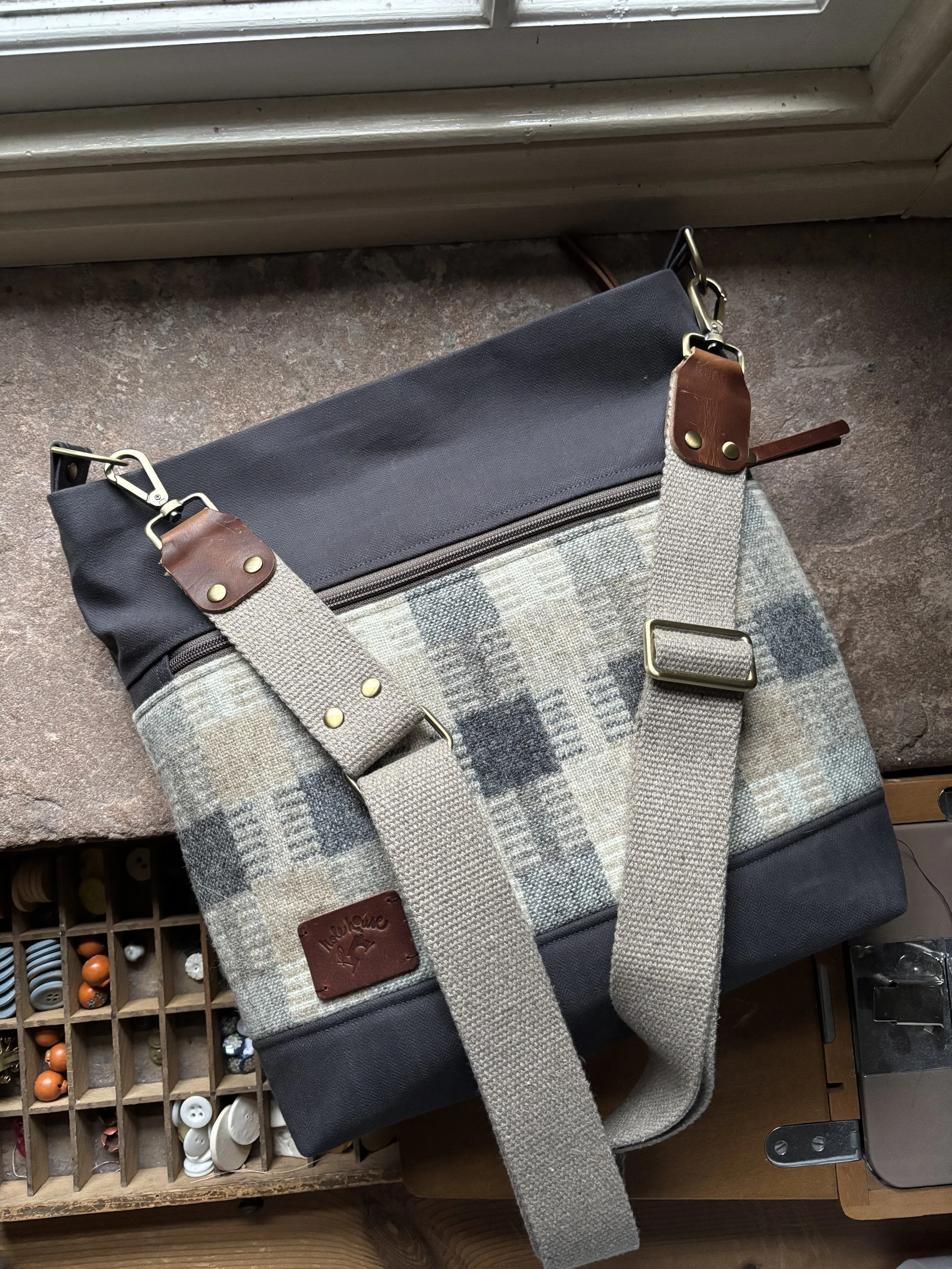 A tote bag with a fabric patterned center, leather accents, and adjustable straps, placed on a wooden surface near a window.