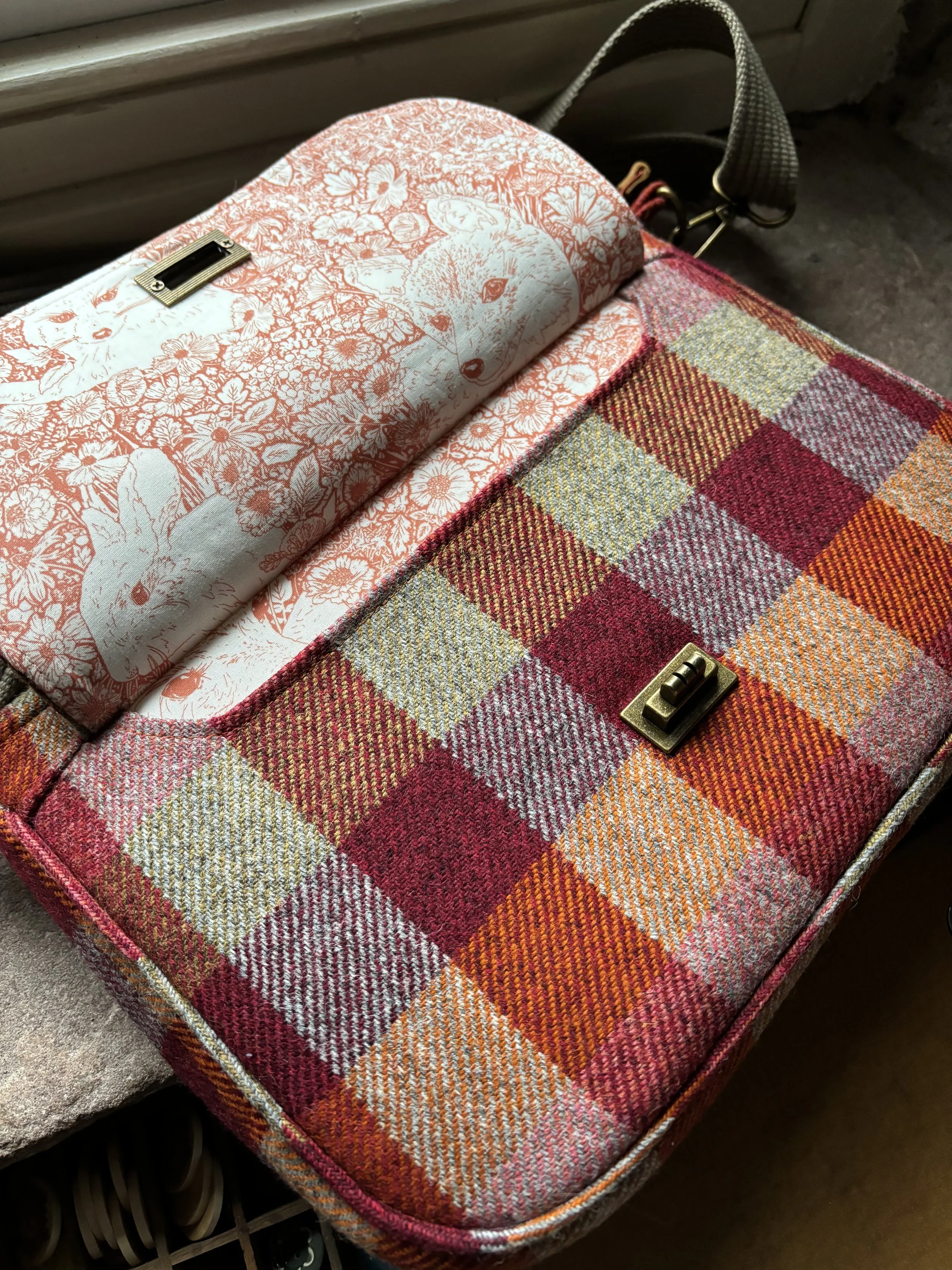 A rectangular plaid fabric handbag with a gold clasp on top, placed on a surface near a window. The bag features a red, orange, yellow, and pink checkered pattern. A folded cloth with rabbit and floral illustrations in red and white rests on top of the bag.