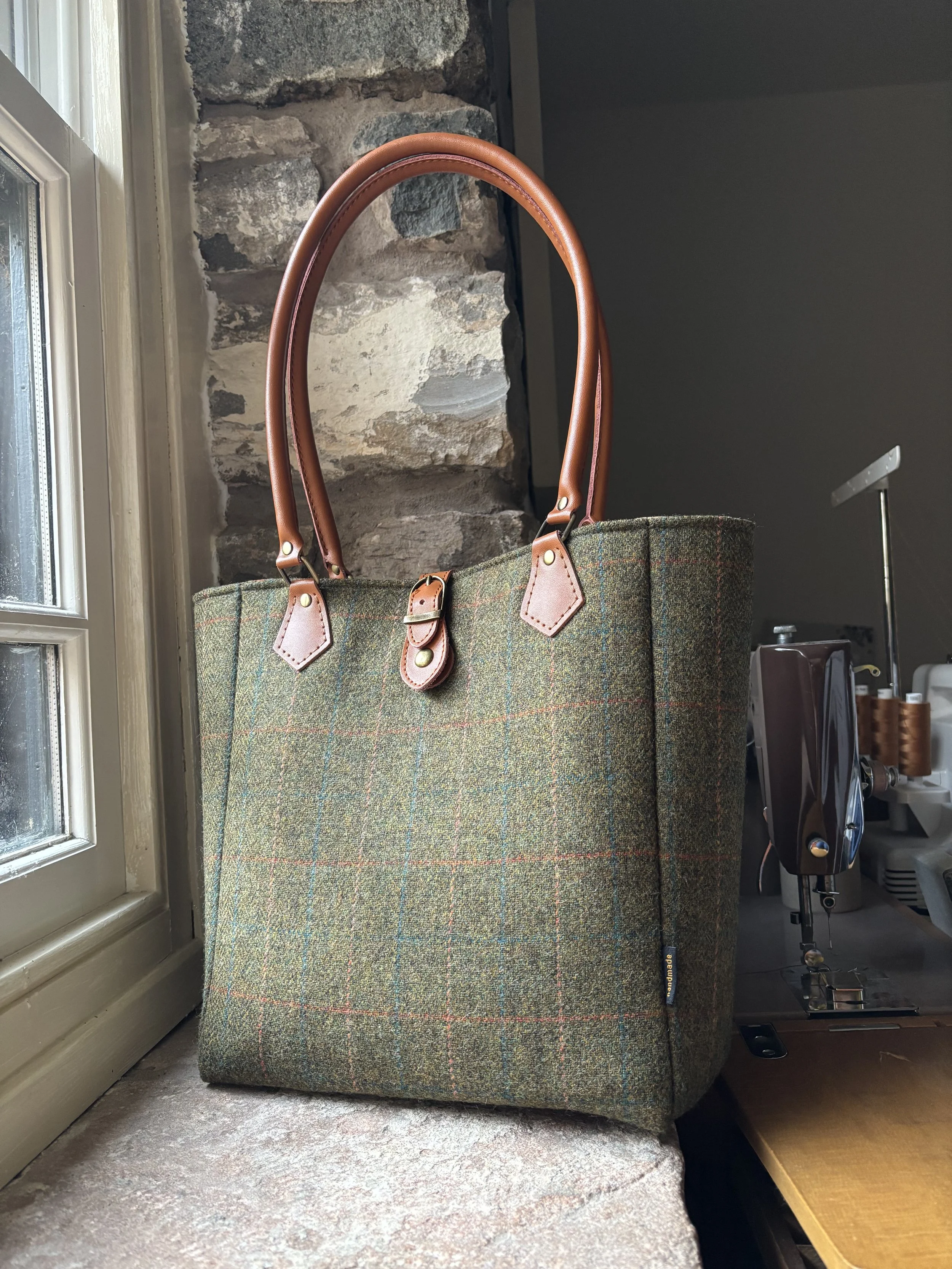 A large tote bag made of green plaid wool with leather handles and accents, placed on a windowsill with a stone wall in the background.