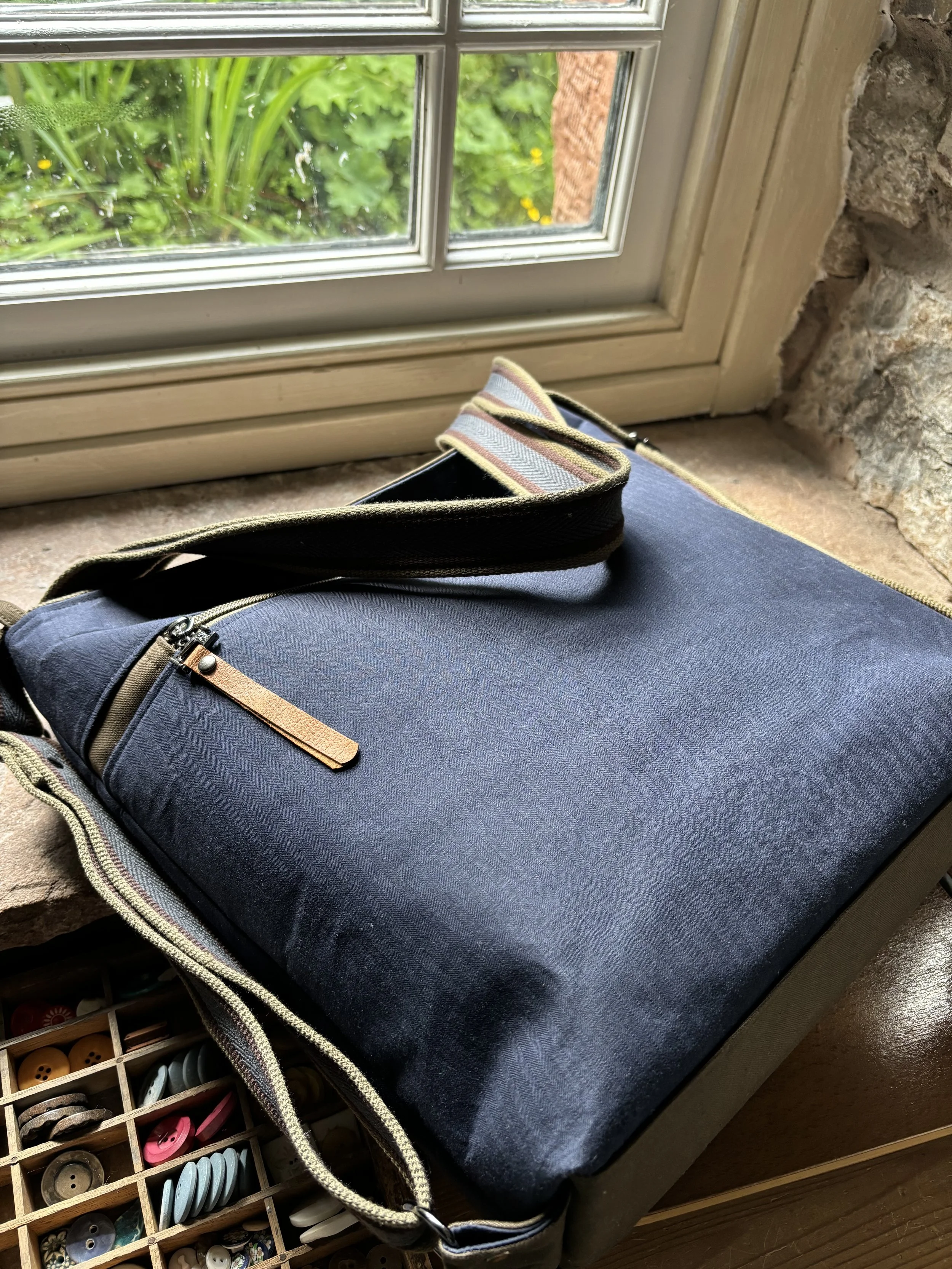 A closed dark gray laptop bag with a leather zipper pull, resting on a wooden surface near a window overlooking greenery outside.