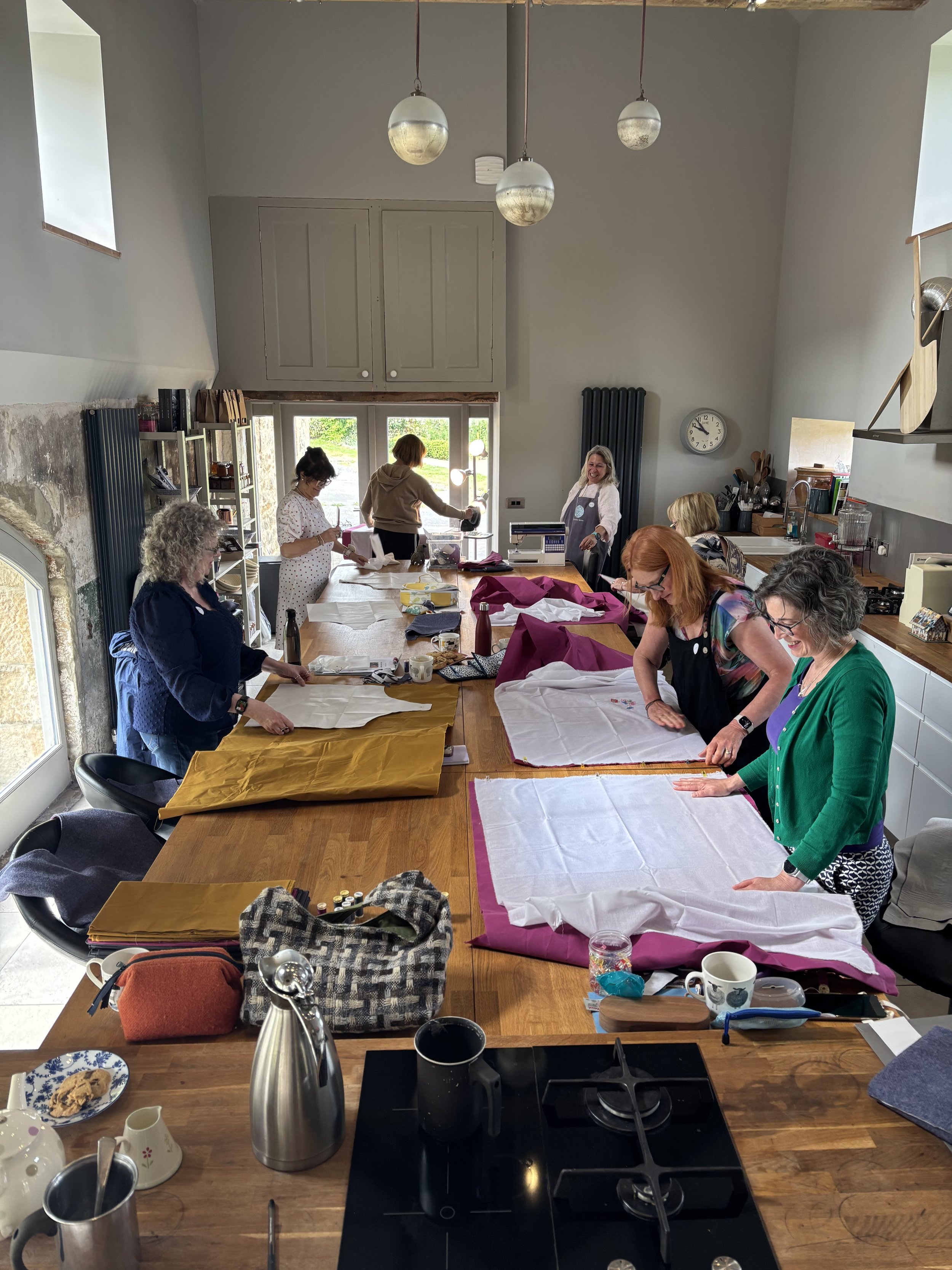 A group of women working on fabric projects around a long wooden table in a bright kitchen, with supplies scattered on the table.