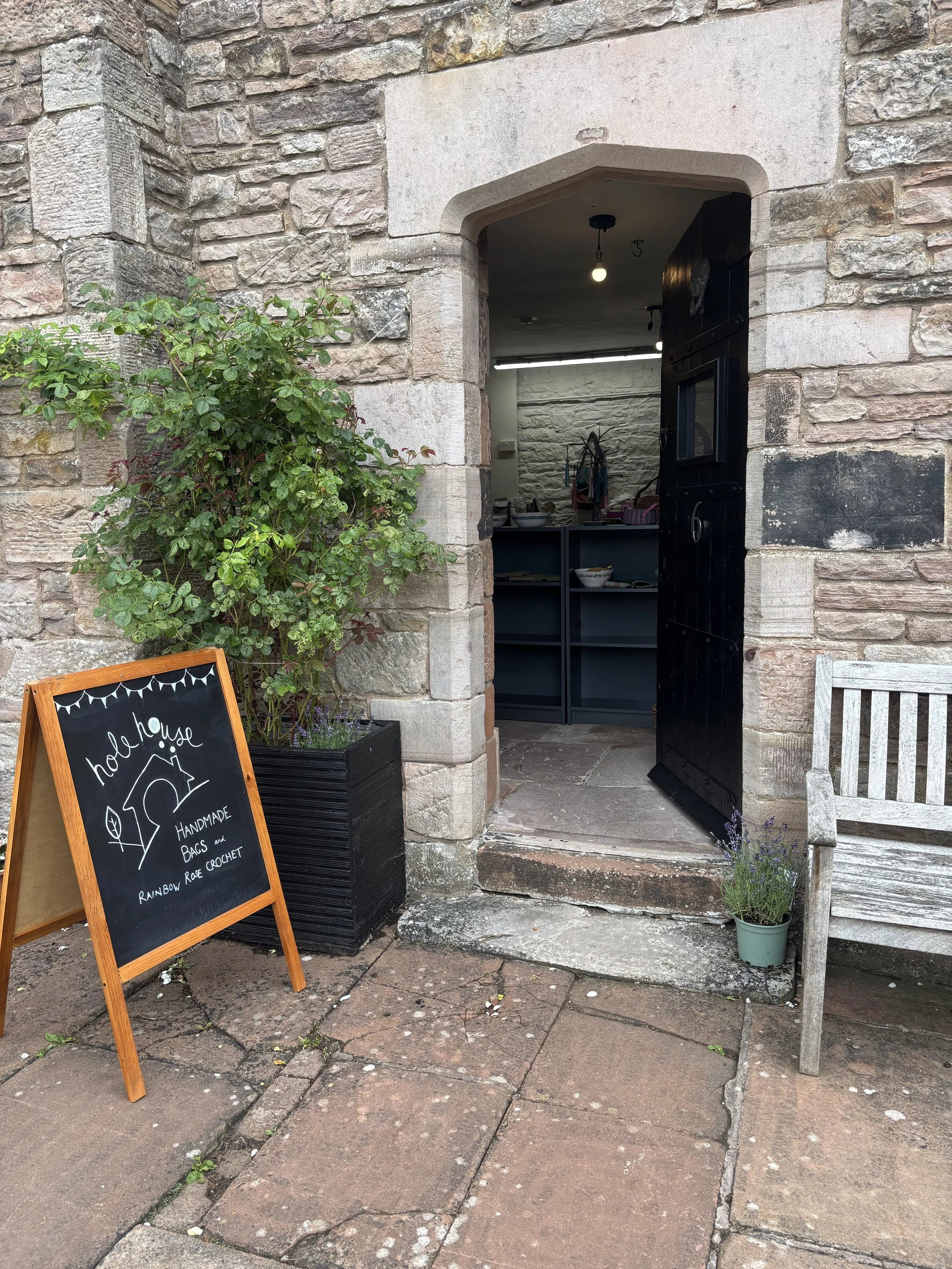 The entrance of a shop with a black door, a chalkboard sign advertising handmade bags and rainbow rose crochet, a large green shrub in a black planter, and a wooden bench outside. The interior appears to have shelves and a counter.