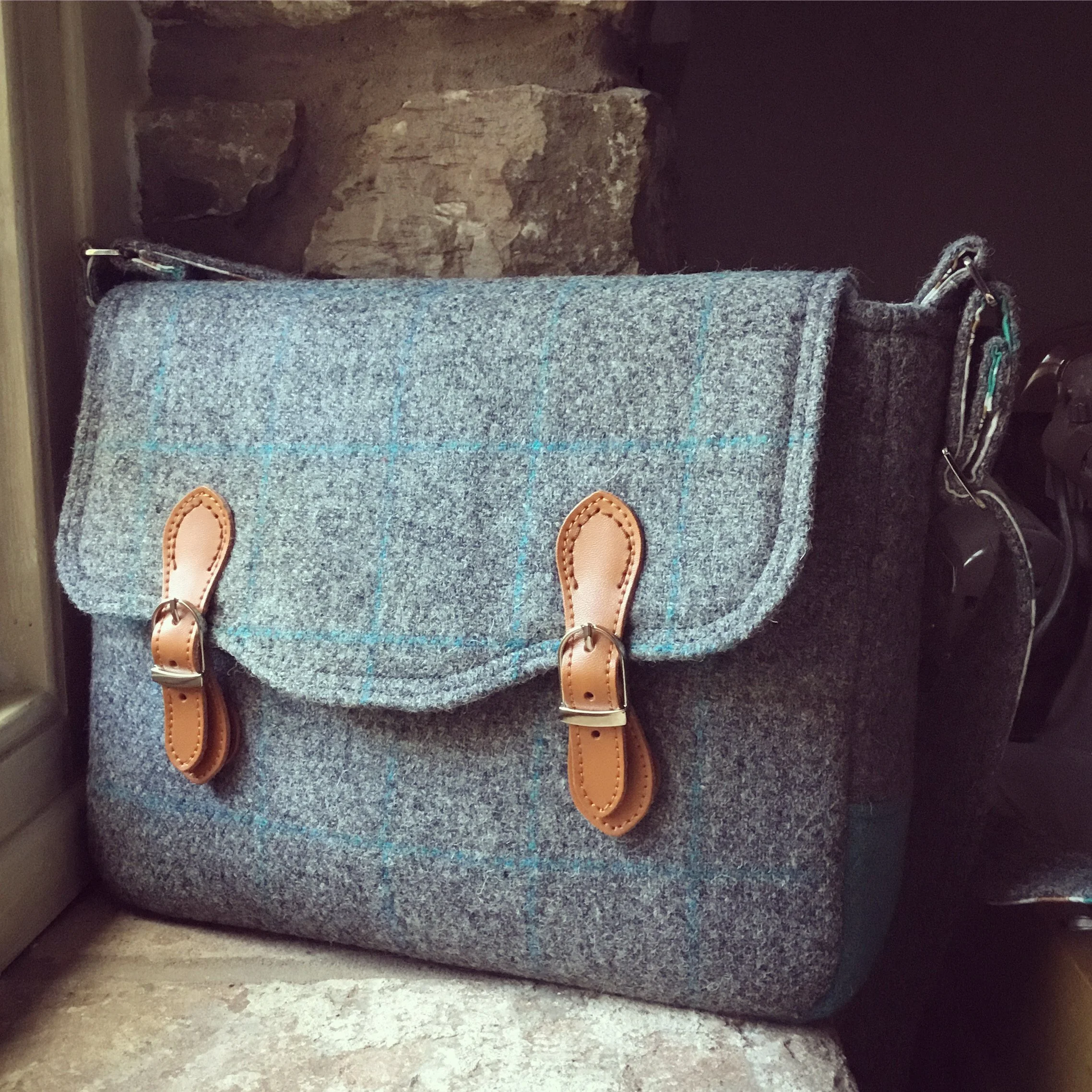 A gray plaid fabric shoulder bag with tan leather straps and buckles, resting on a stone surface near a stone wall.