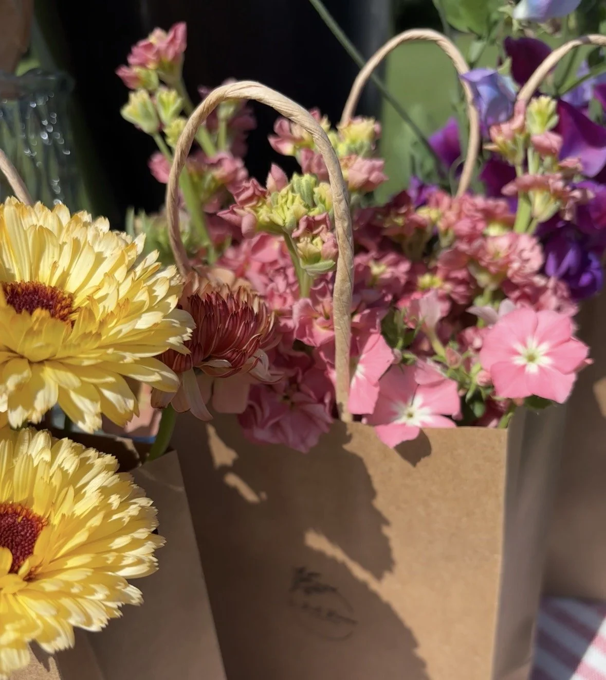 A paper bag filled with a colorful variety of fresh flowers, including yellow daisies, pink delphiniums, and purple petunias, set outdoors.
