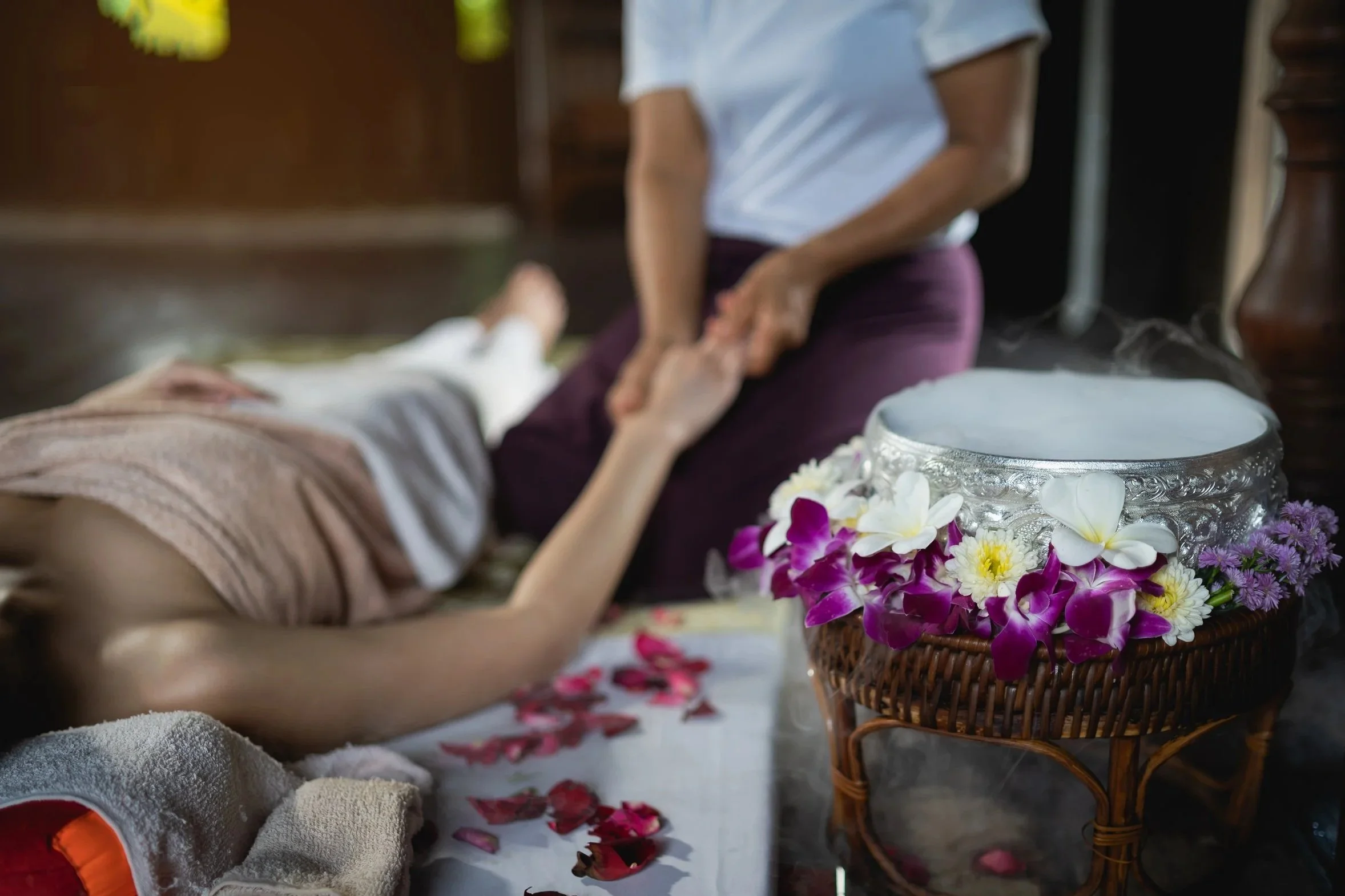 A person lying on a massage table receives a traditional Thai herbal compress massage in a spa setting. The person’s legs are visible, and they are covered with a towel. There is a steaming herbal compress and flowers nearby.
