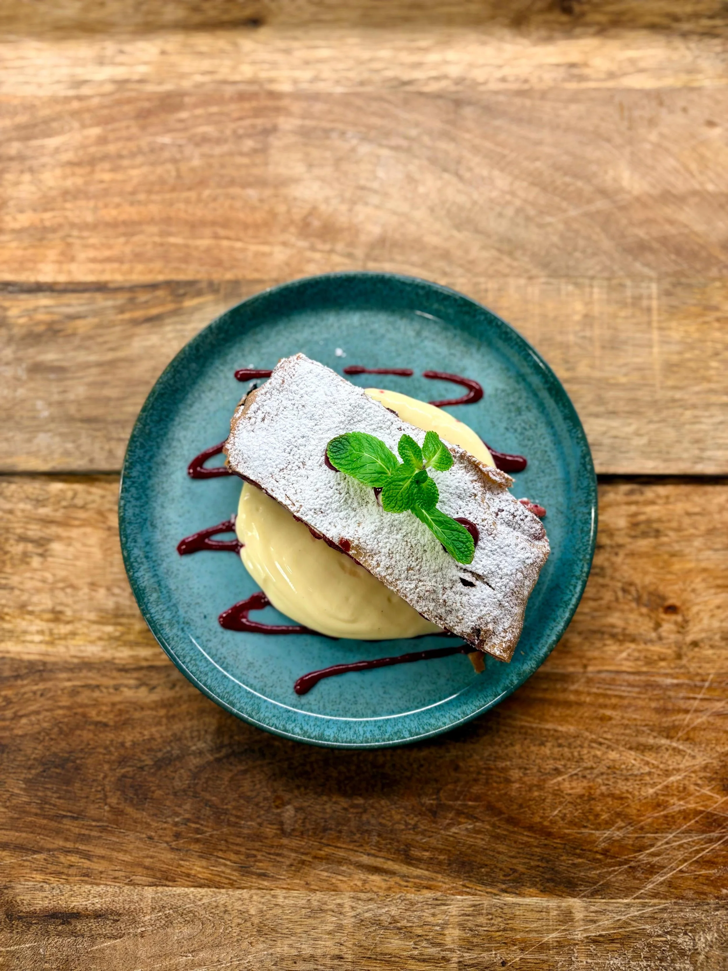 Essen im Martinus in Weingarten: Ein Kuchen mit einer Schicht Vanillecreme, garniert mit einem Minzblatt, auf einem blauen Teller mit Schokoladensauce, auf einem Holztisch.