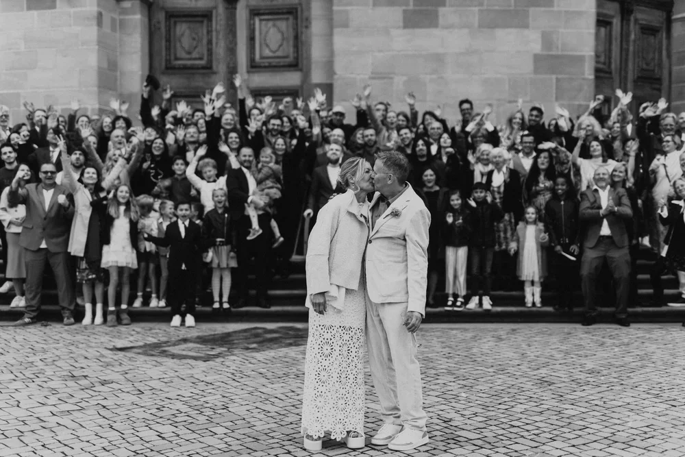 Ein Paar bei einer Hochzeitszeremonie küsst sich vor den Türen des Martinus in Weingarten, während eine große Gruppe von Menschen im Hintergrund zusieht. 
