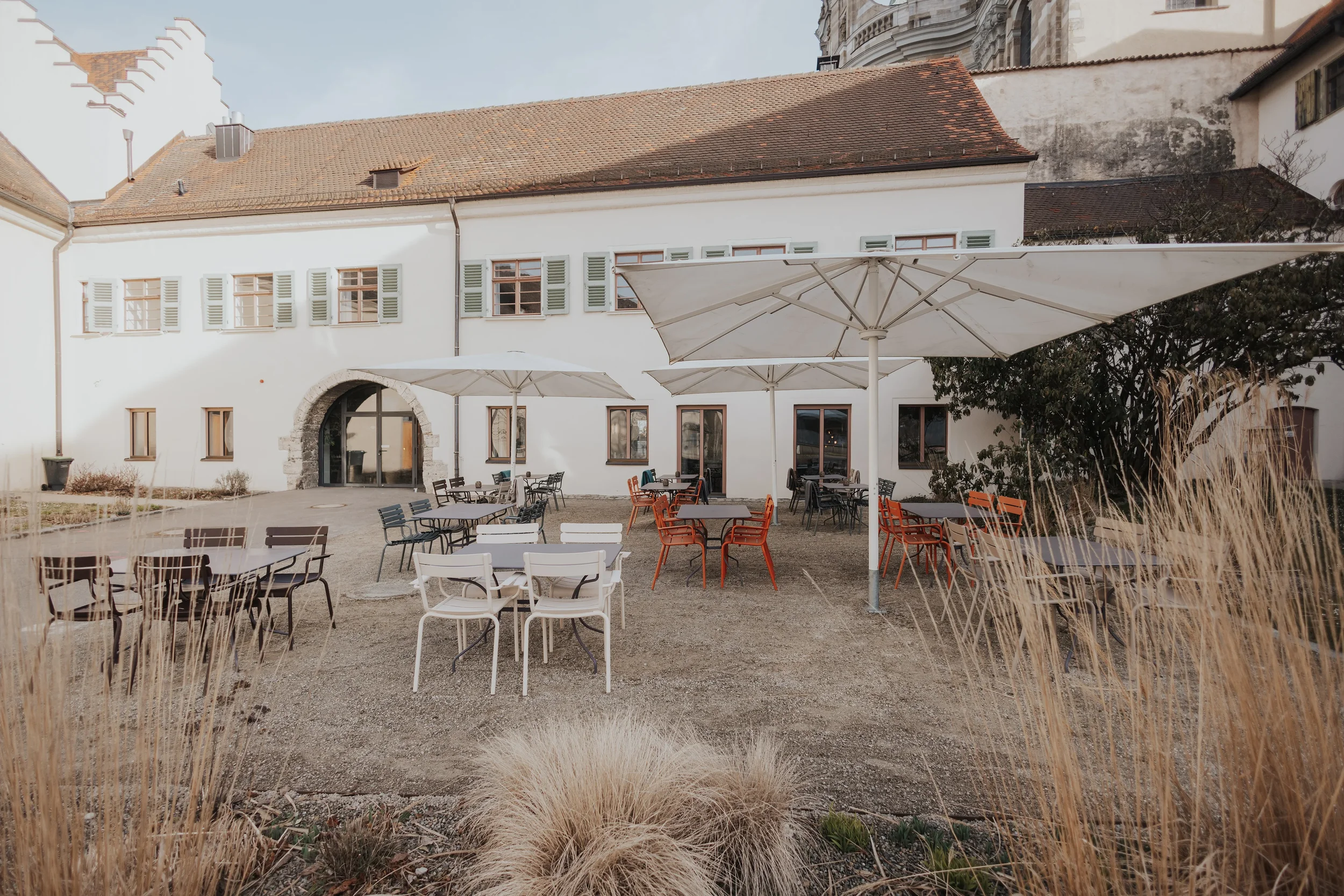 Der Außenbereich des Martinus in Weingarten mit Tischen und Stühlen, großen Sonnenschirmen und einem weißen Gebäude im Hintergrund.