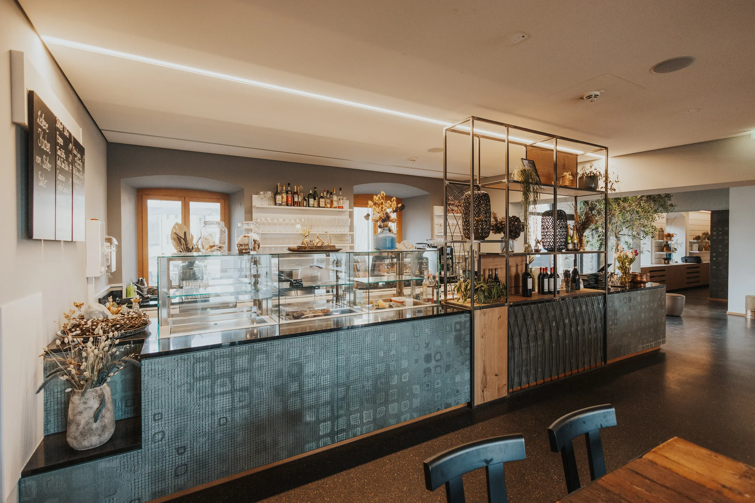 Blick auf die moderne, gemütliche Café-Bar mit Glasvitrine, darin Süßigkeiten und Gebäck, und einer dekorativen Regalwand mit Pflanzen, Flaschen und Dekorationsartikeln. Im Hintergrund sind Sitzbereiche und Fenster vom Martinus sichtbar.