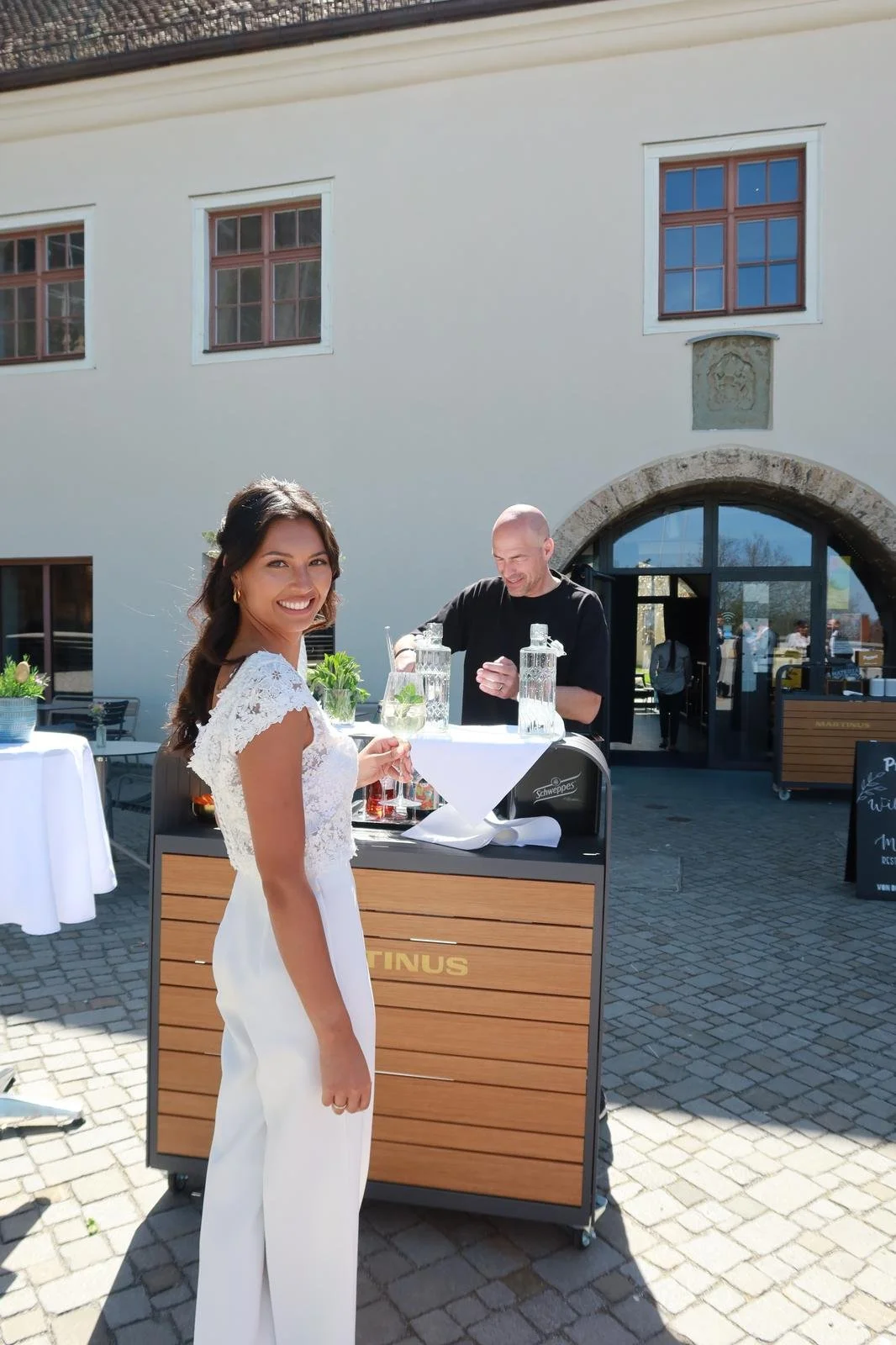 Eine Frau in einem weißen Kleid hält ein Getränk und lächelt, während ein Barkeeper hinter einem mobilen Bar-Stand Getränke zubereitet, im Freien vor dem Martinus in Weingarten.