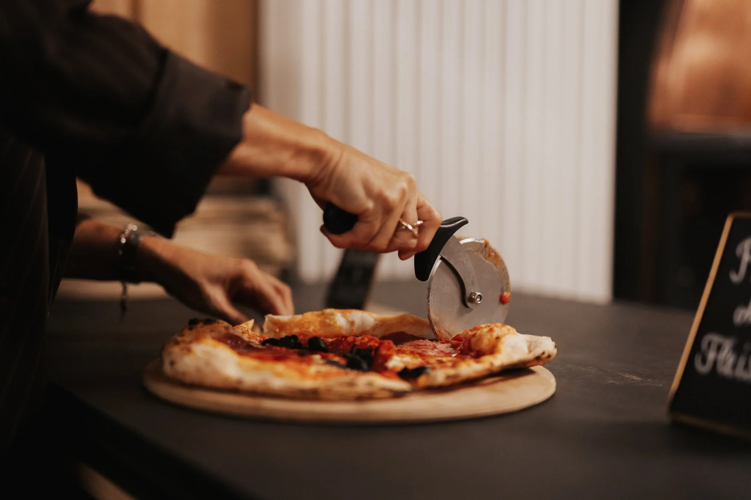 Person schneidet eine Pizza mit einem Pizzaschneider auf einem Holzbrett.