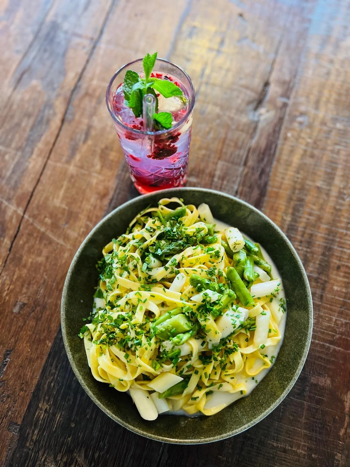 Essen im Martinus in Weingarten: Nudelsalat mit grünen Bohnen und Kräutern auf einem grünen Teller, daneben ein Glas mit rosa Getränk, Minzblättern und Eis, auf Holzoberfläche.
