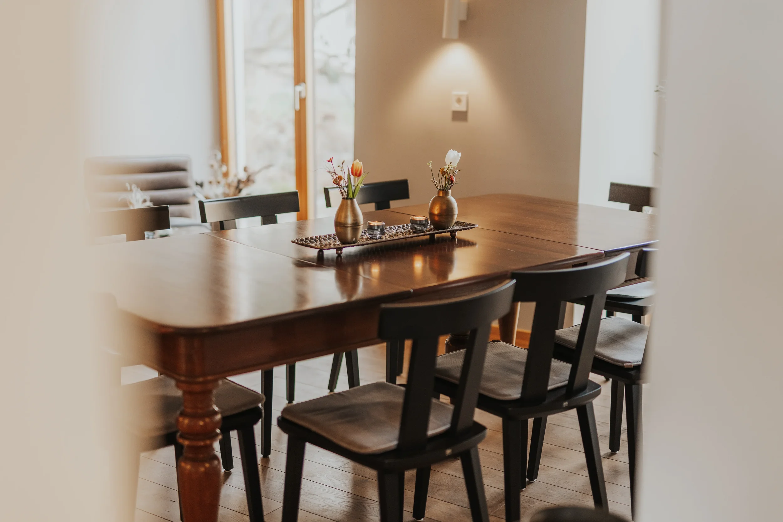 Eleganter Essbereich mit dunklem Holztisch, schwarzen Stühlen, zwei bronze Vasen mit Blumen, in einem hellen Raum mit großen Fenstern.