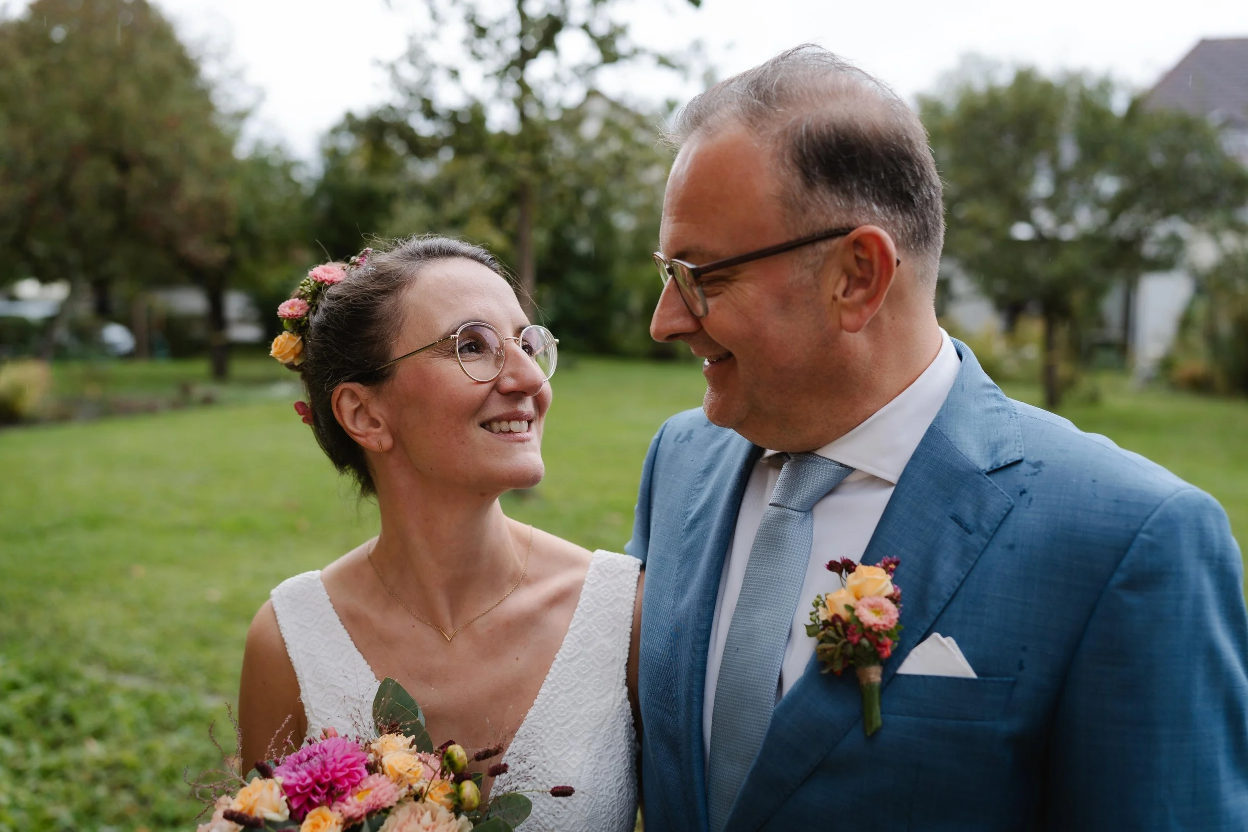 Ein glückliches Brautpaar bei einer Hochzeit im Freien, umgeben von Bäumen und grünem Rasen, lächelnd und Blickkontakt.