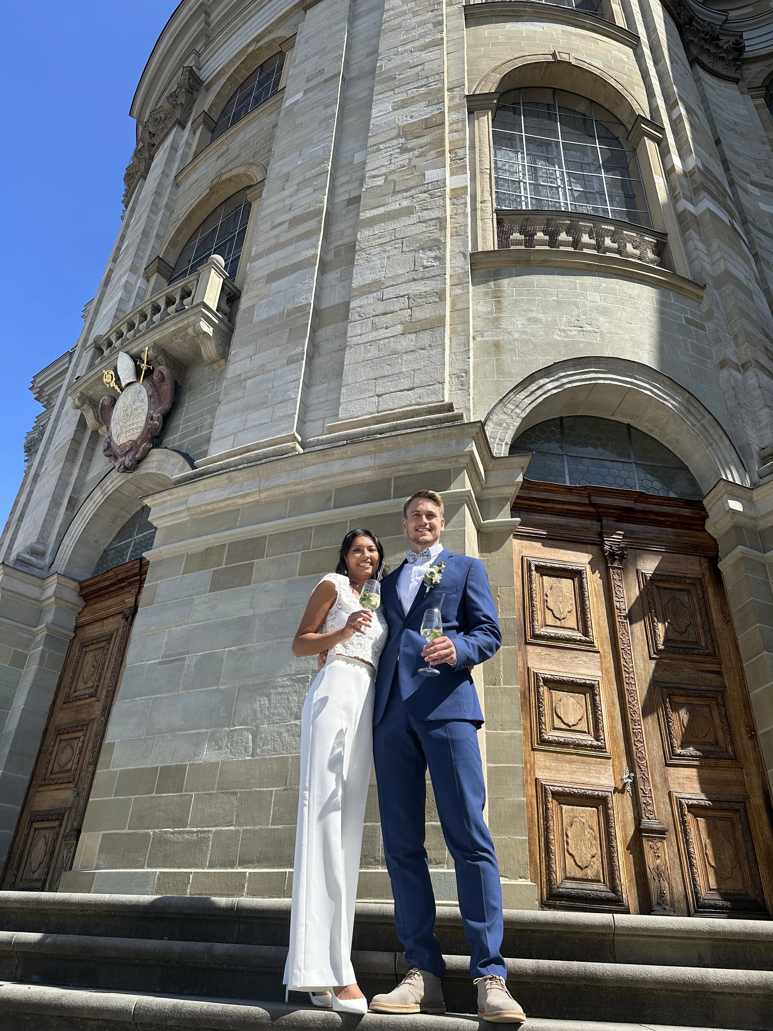 Ein frisch vermähltes Paar steht vor einer historischen Kirche, sie trinken Sekt, Sonnenschein, festliche Stimmung.